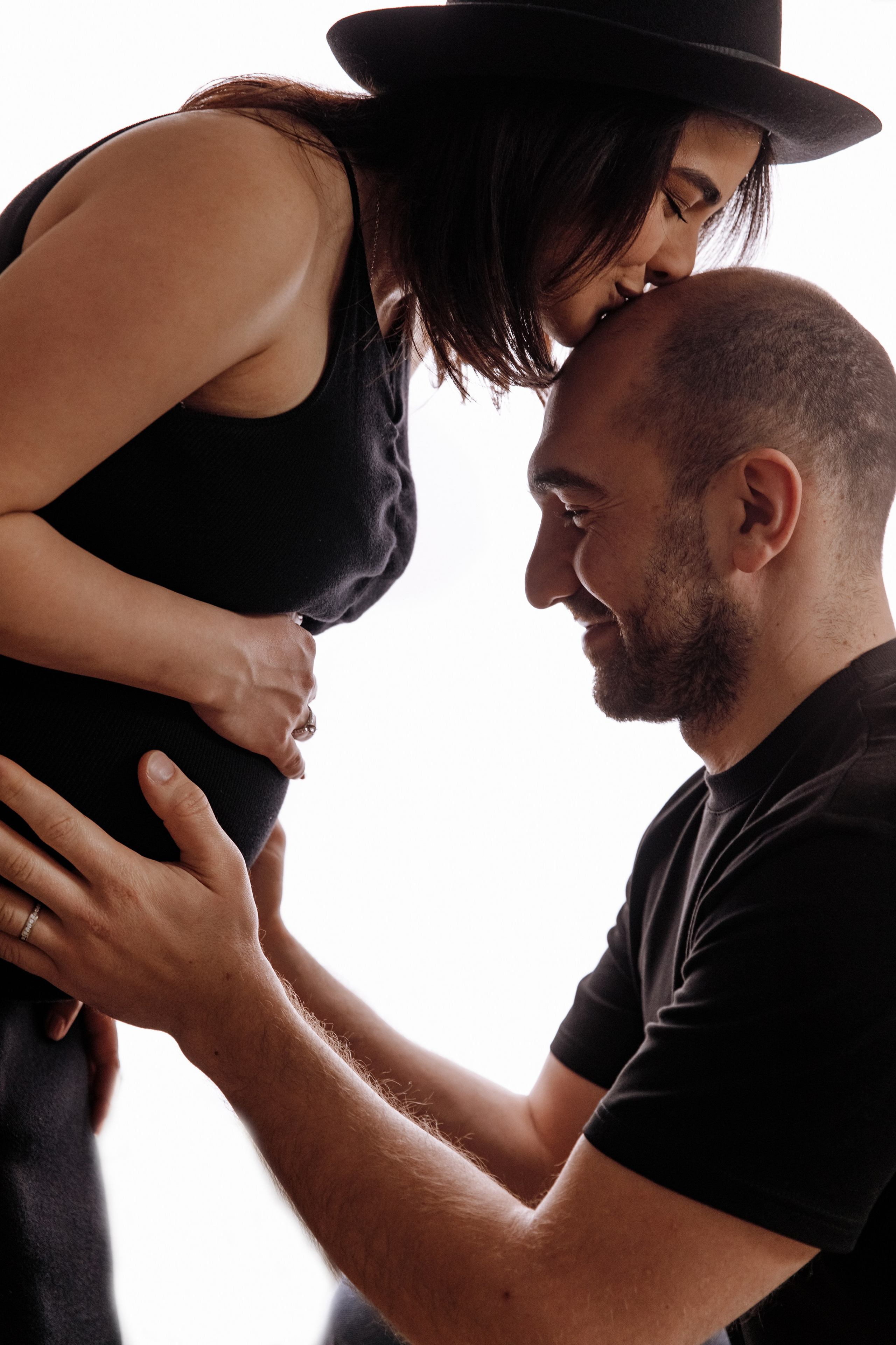 Mann hält den Babybauch seiner schwangeren Frau, während sie ihm liebevoll einen Kuss auf die Stirn gibt.