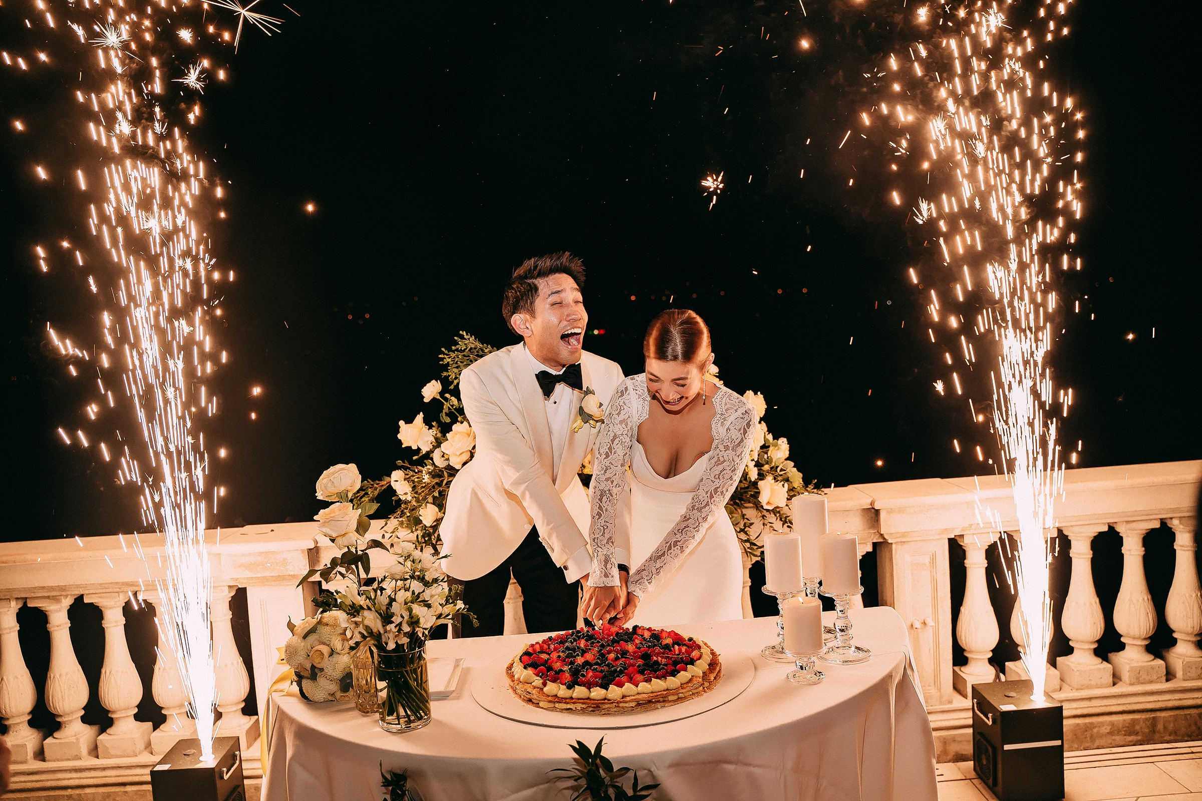 A bride and groom joyfully cut their wedding cake with sparklers illuminating the night behind them.