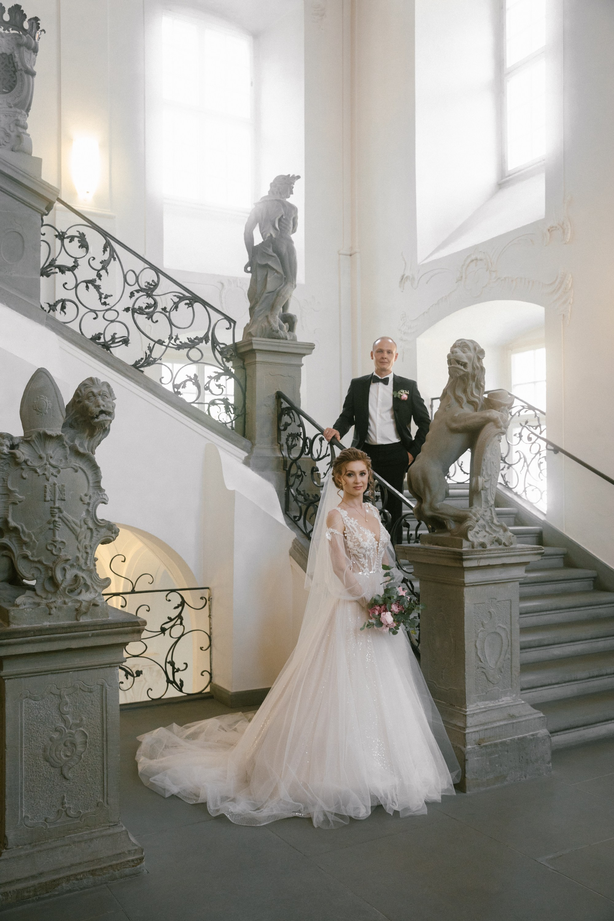 Bride stands in front of staircase holding bouquet, groom above on steps at Schloss Meersburg