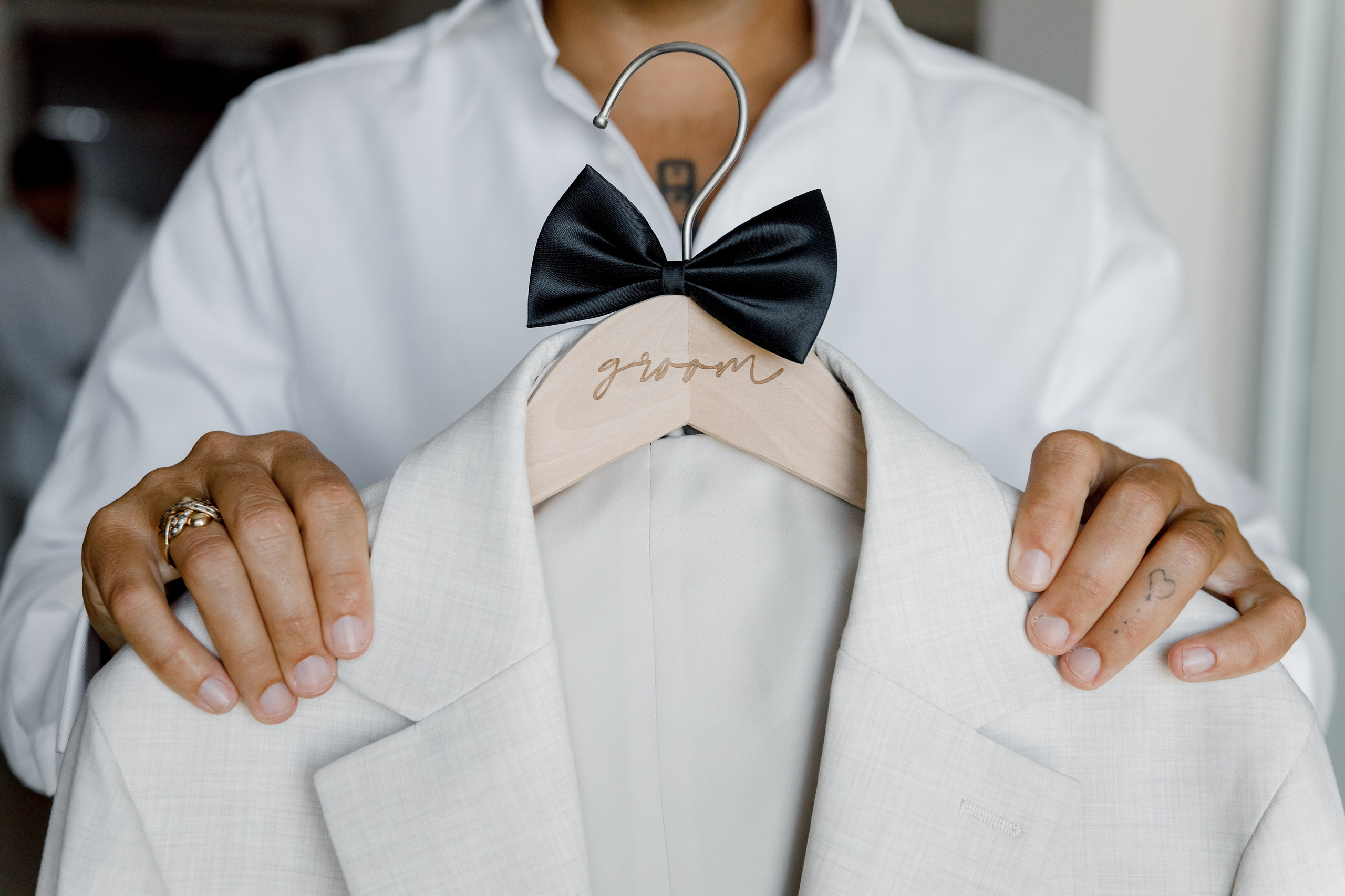 The groom stands confidently in a crisp white dress shirt, holding his tailored biege suit on a wooden hanger engraved with ‘The Groom’ in elegant script.
