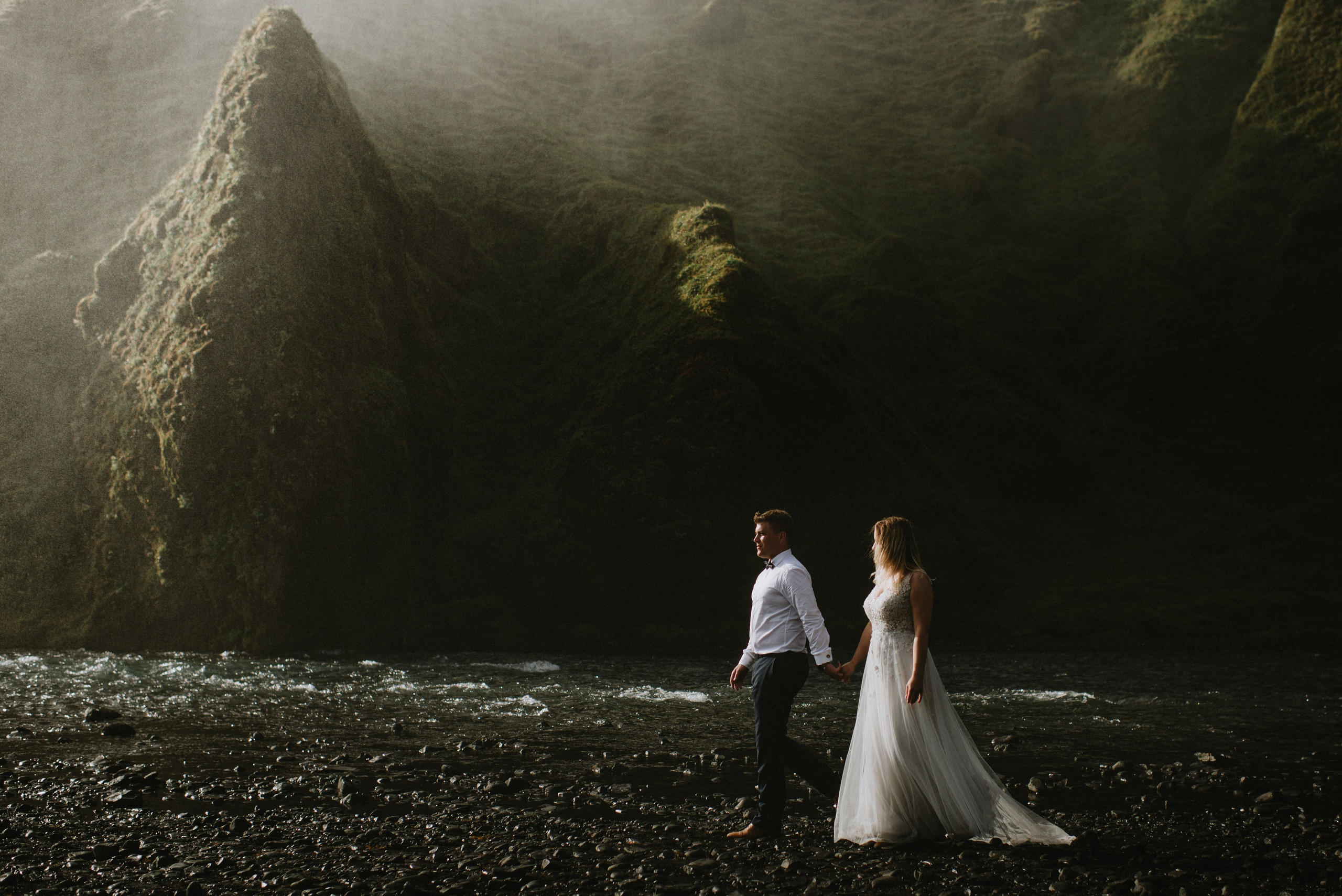 Bride and groom looking up at the towering Skógafoss waterfall, holding hands.