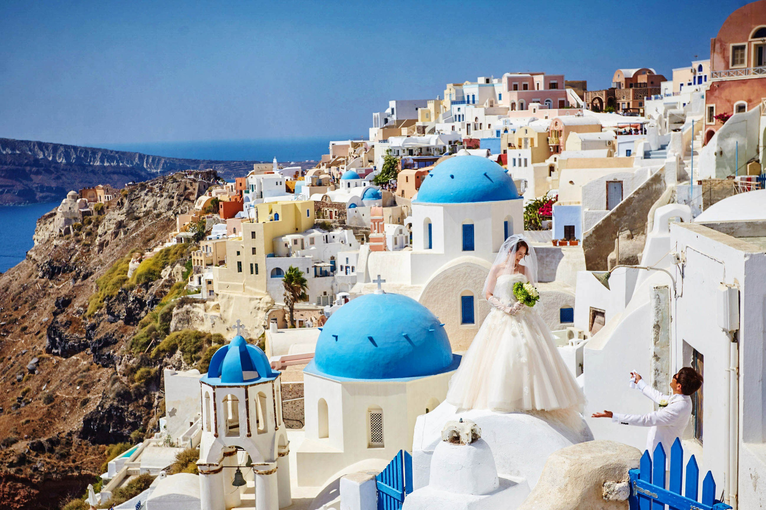 A groom tries to get the brides attention as she stands atop the white roofs of Santorini during her portrait session.