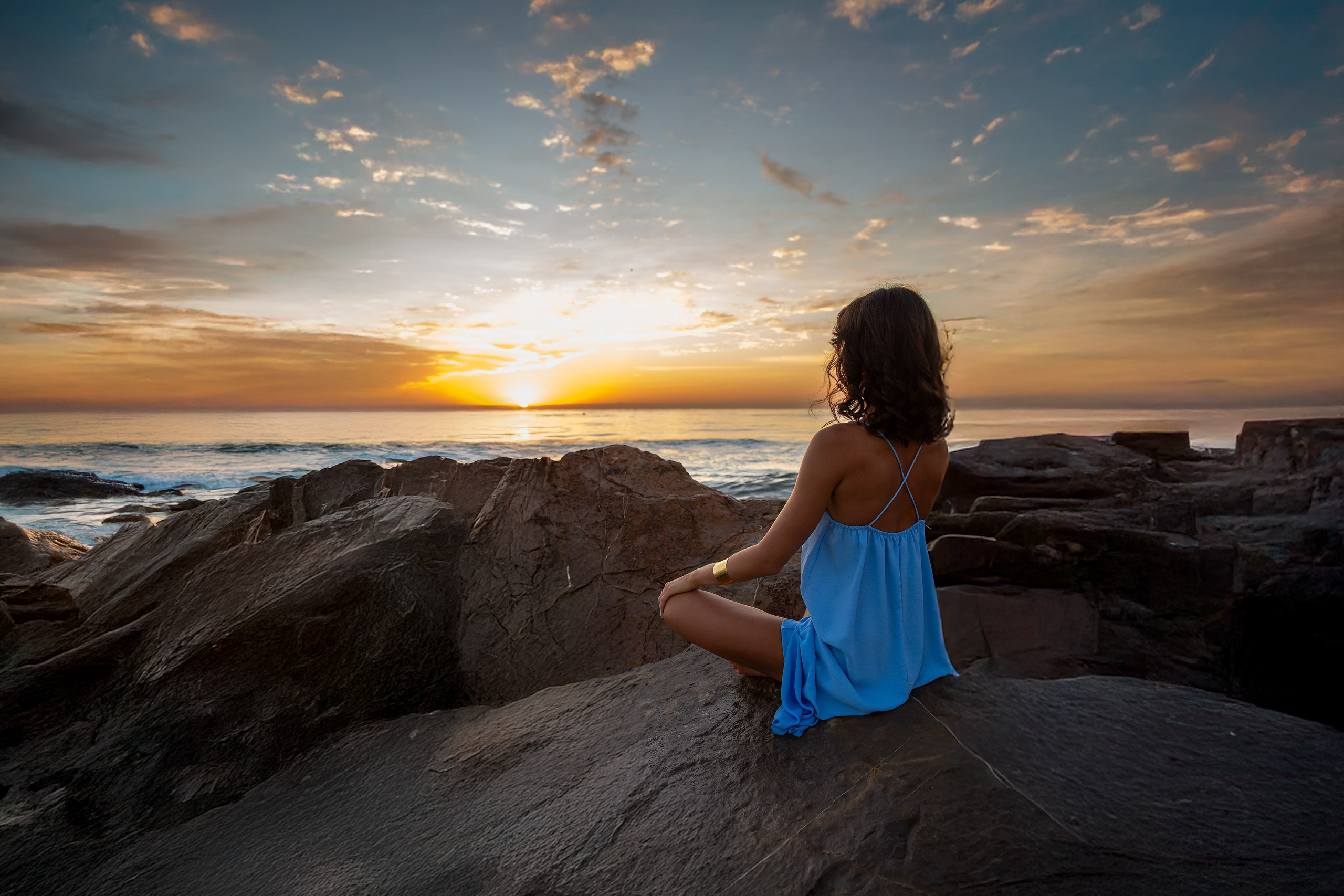 Спокойная фотосессия на закате в Дении, Испания — девушка в синем платье сидит на прибрежных скалах, глядя на золотой горизонт над Средиземным морем. Отличный выбор для тех, кто ищет умиротворённую и художественную портретную съёмку в Дении и вдоль Коста-Бланки.