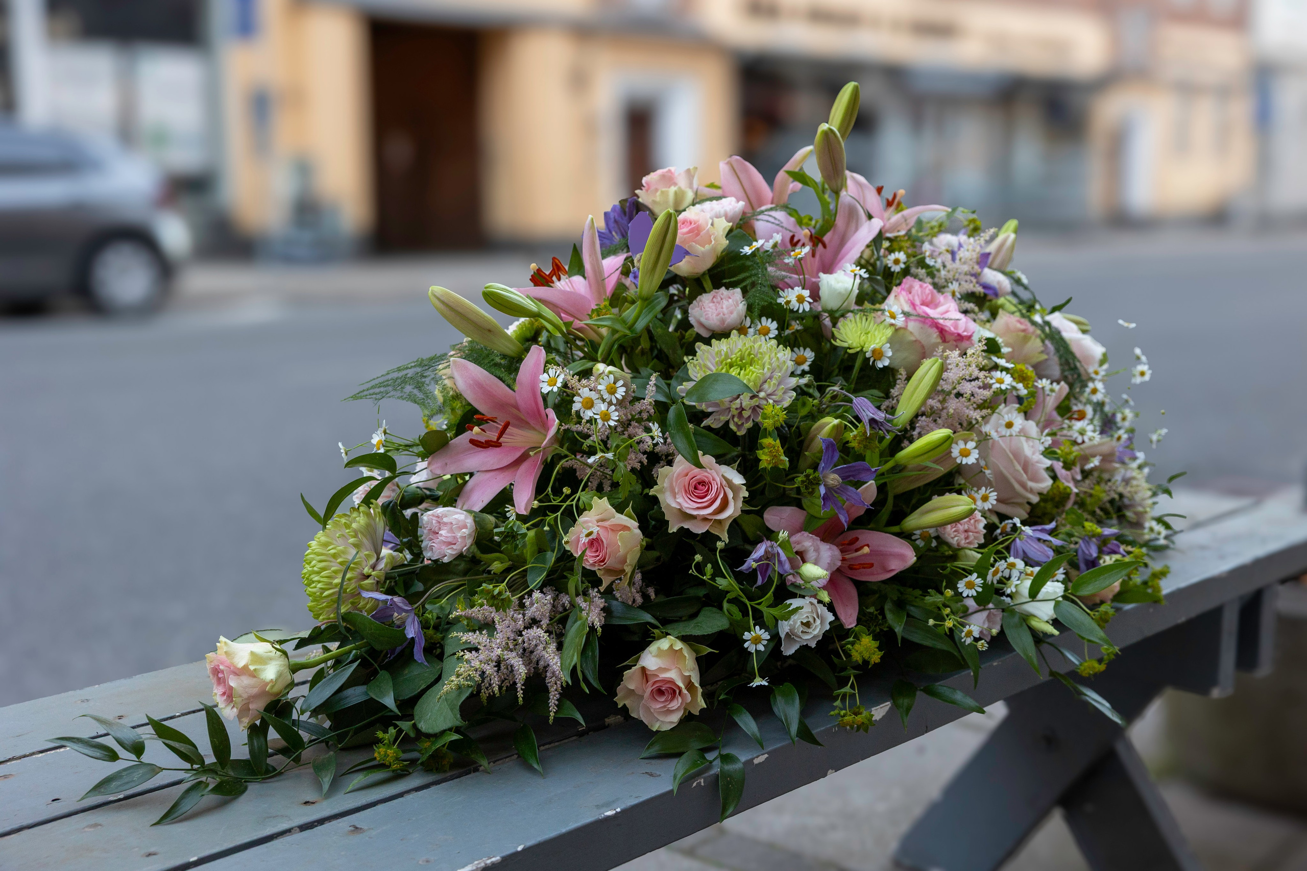 Denne luftige kistedekoration er sammensat af liljer, roser og små blomster i bløde pastelfarver. Akropolis Blomster skaber unikke begravelsesblomster til kirker, kapeller og bisættelser i Aarhus.