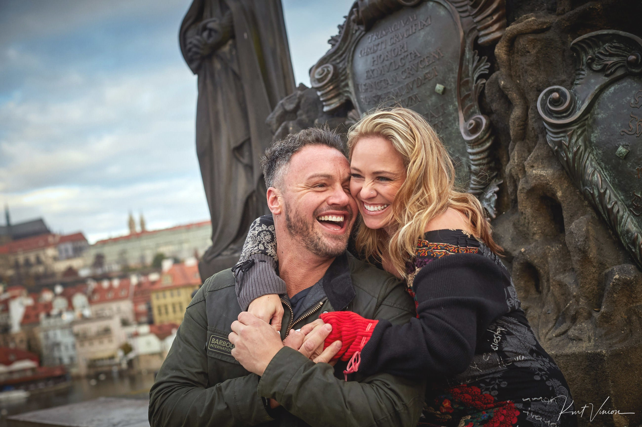 Woman embracing man atop Charles Bridge smiling before marriage proposal in Prague.