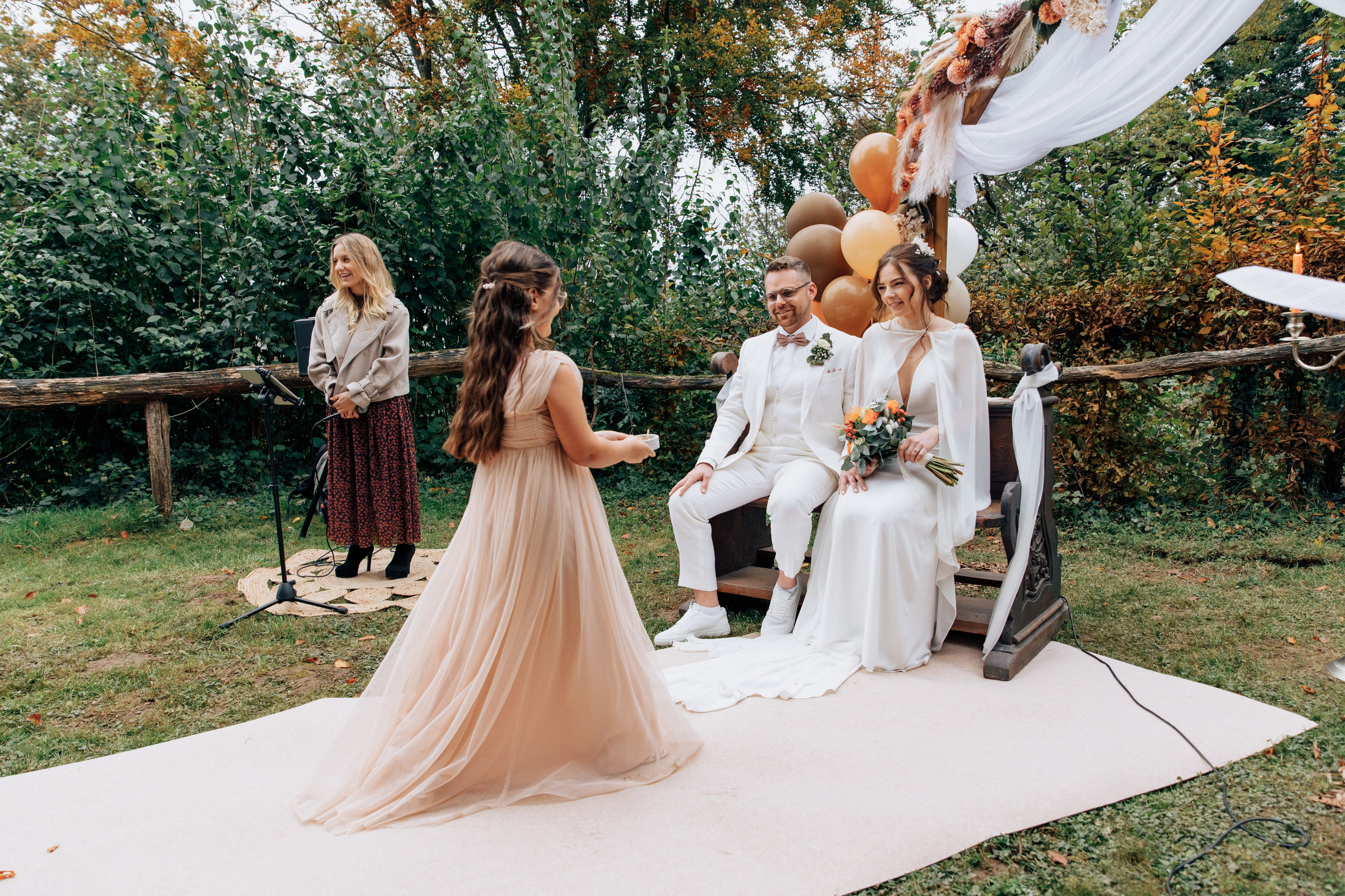 Ein Mädchen in einem pfirsichfarbenen langen Kleid trägt die Ringe zum Bräutigam und zur Braut, im Hintergrund lächelt die Sängerin.