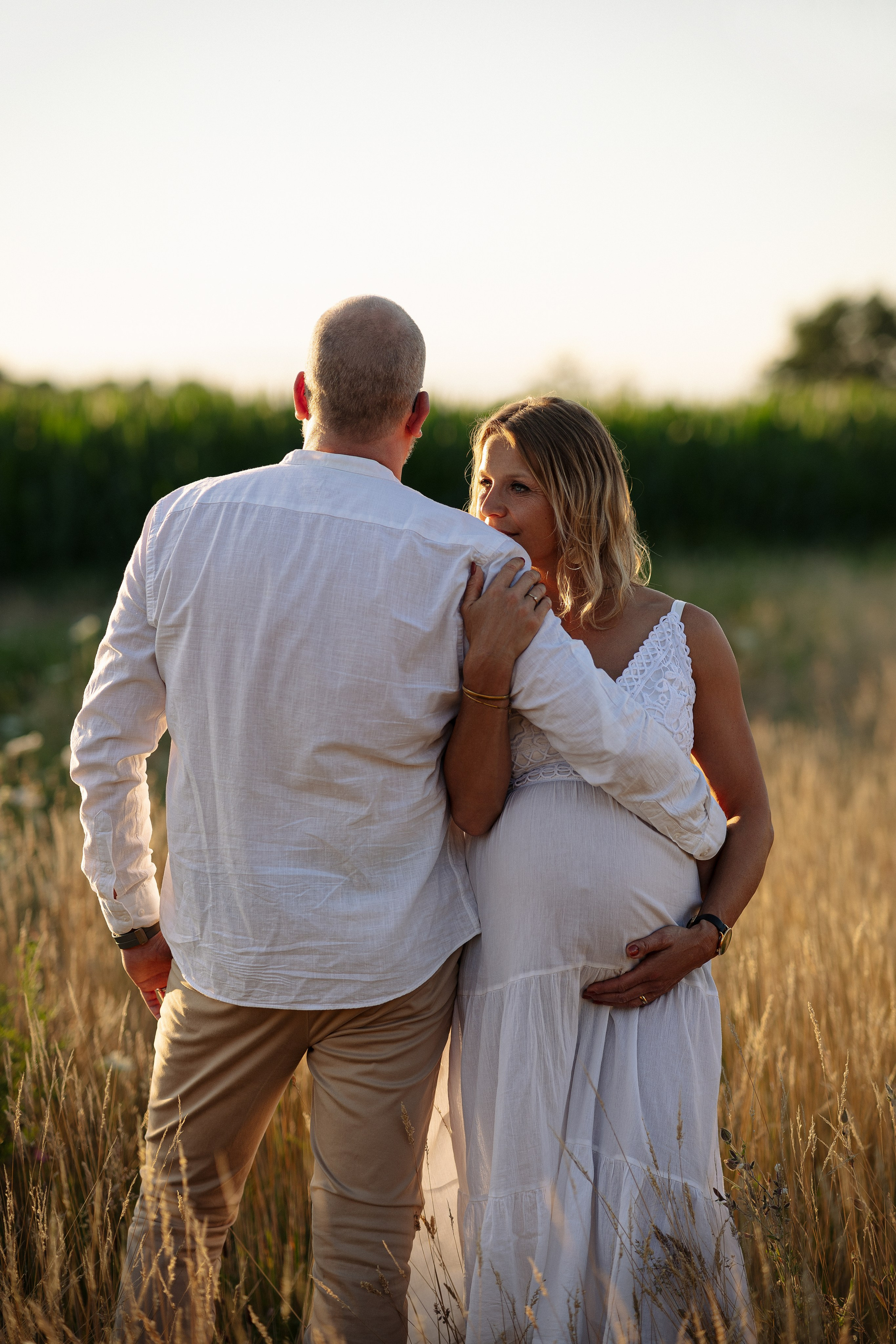 Schwangere Frau umarmt ihren Mann bei Sonnenuntergang während eines romantischen Babybauch Fotoshootings im Feld.