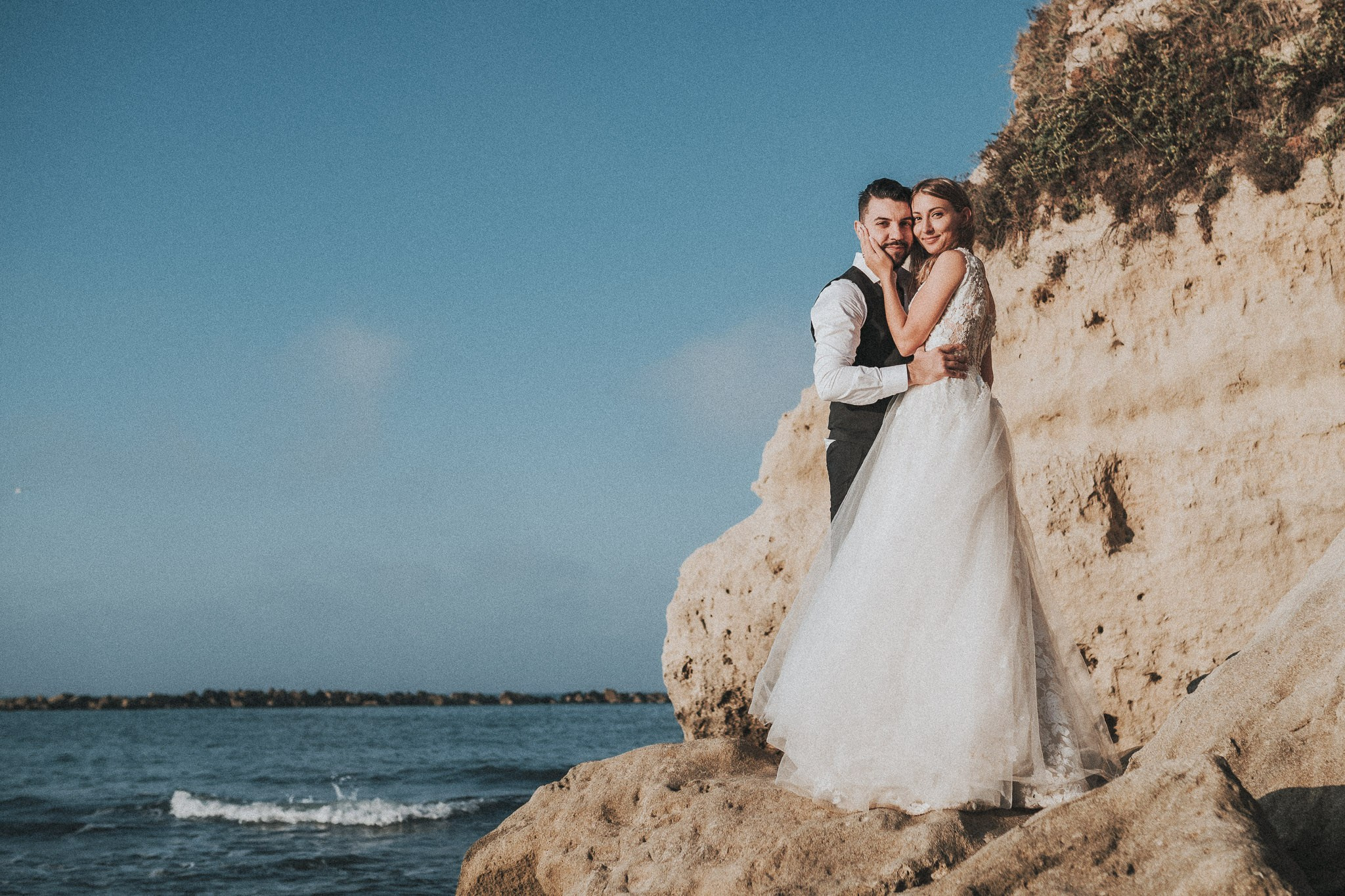 Sposi in posa amorevole su una scogliera, immagine emblematica dello stile unico di un fotografo di matrimonio, con l'oceano che sfuma all'orizzonte.