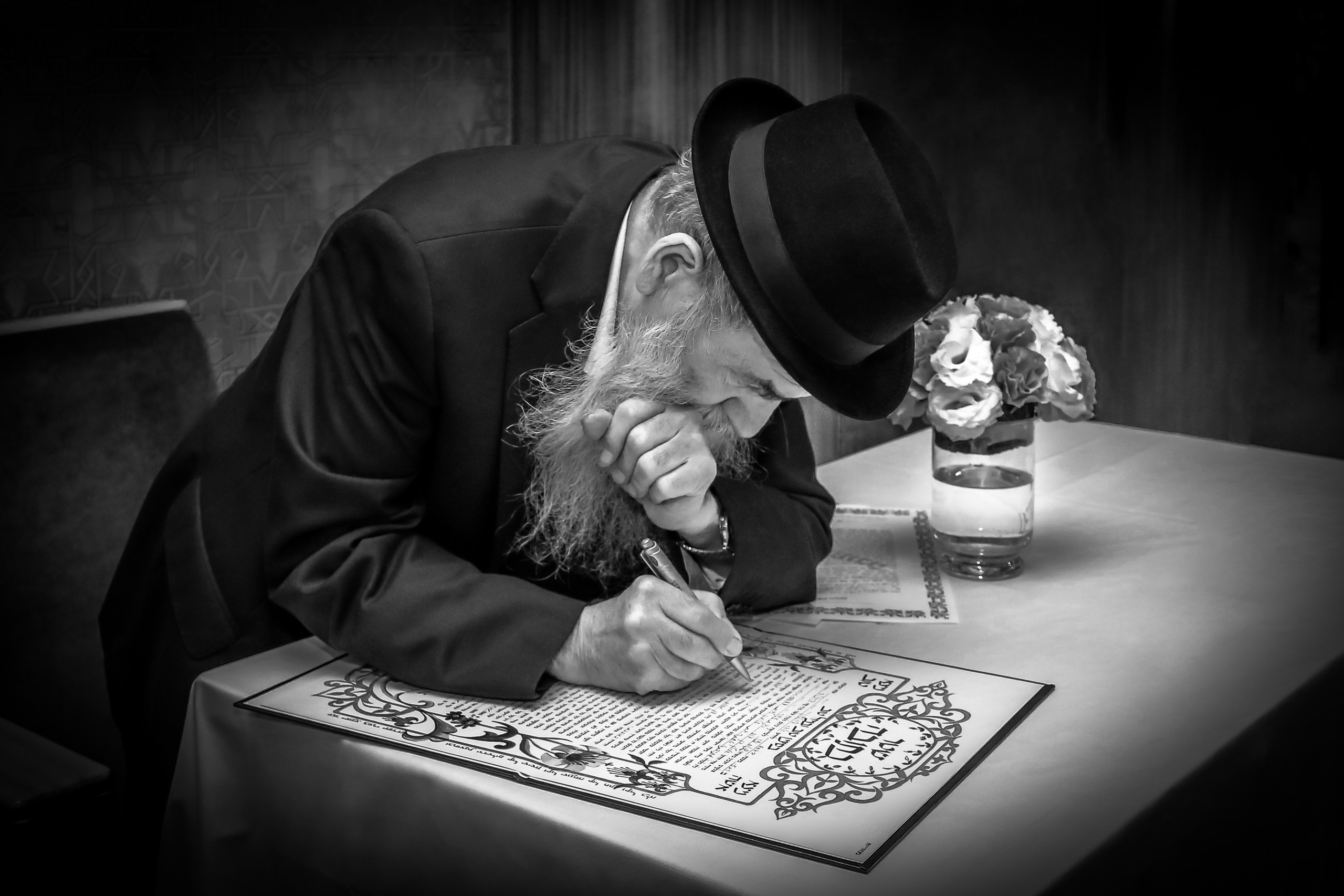 A black-and-white image of a rabbi in a black hat and attire, with a long gray beard, writing the Ketubah, a traditional Jewish wedding contract, at a white table.