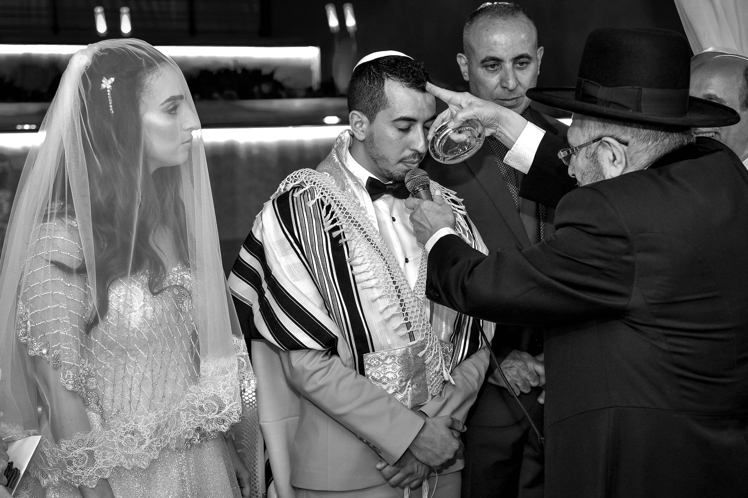 Jewish wedding ceremony under chuppah: Rabbi blesses groom as veiled bride looks on.