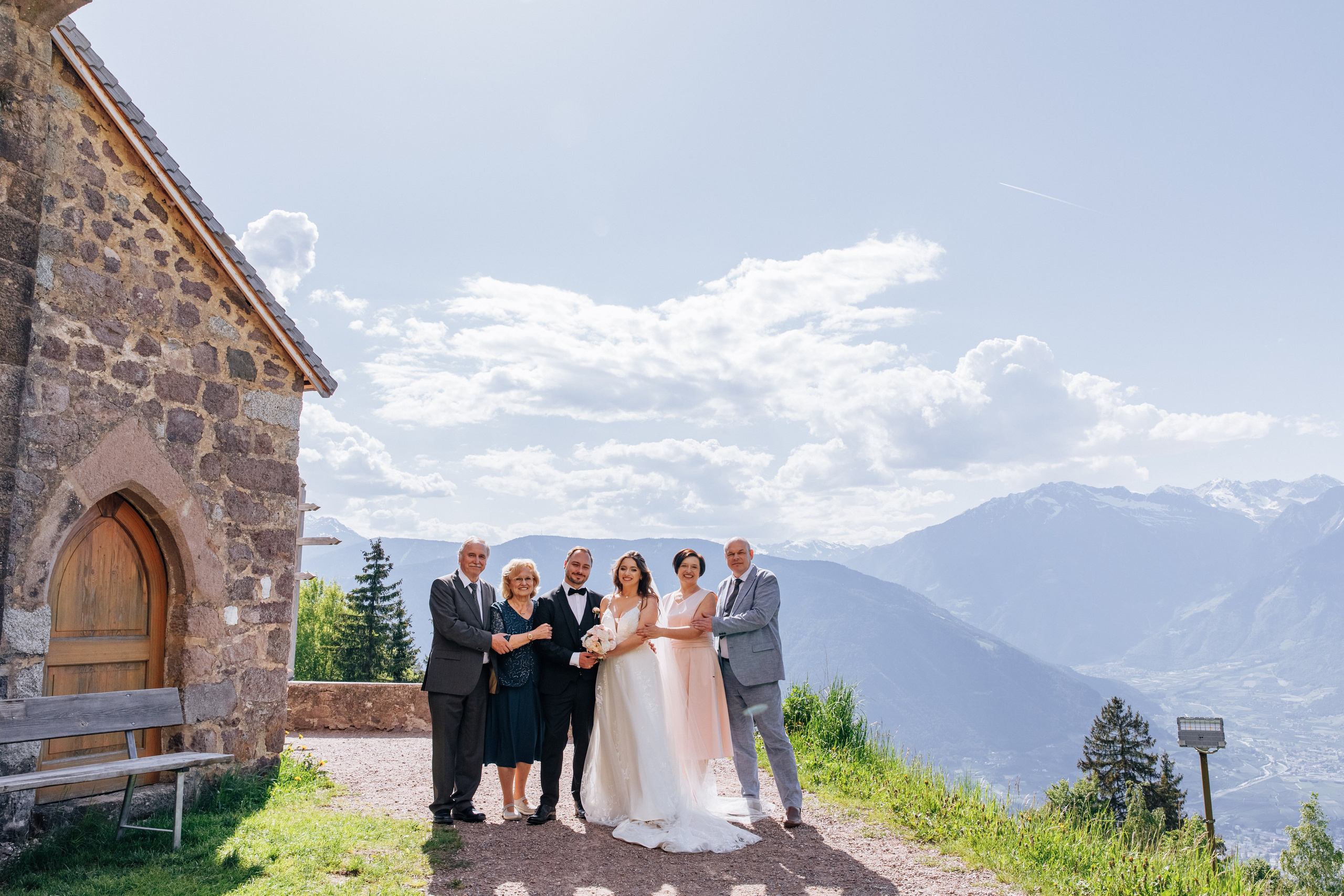 Das Brautpaar steht mit ihren Eltern vor der Kirche und den Bergen als Hintergrund.