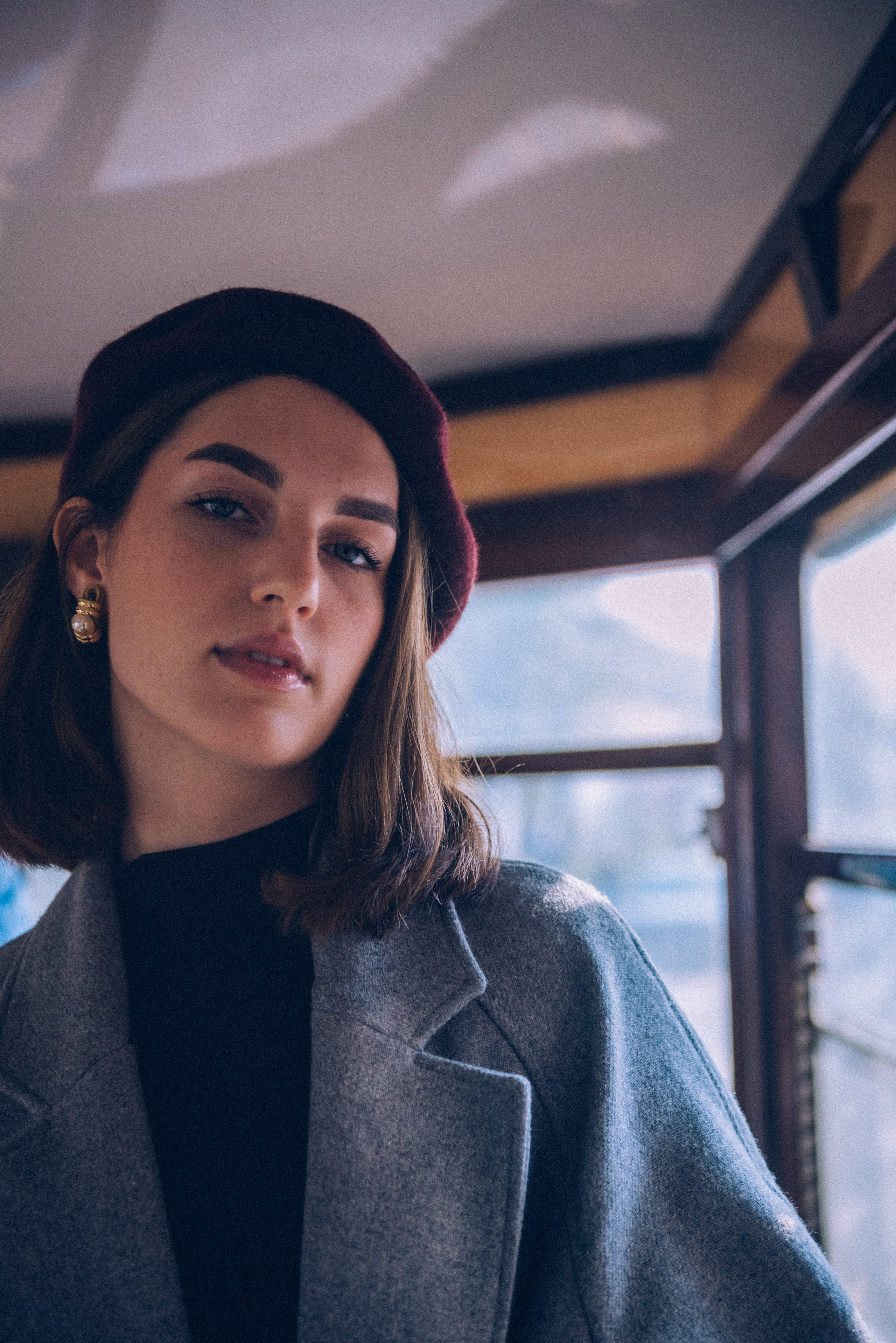 Portrait of a sophisticated woman in a burgundy beret and gray coat, illuminated by soft natural light inside a vintage Milanese tram