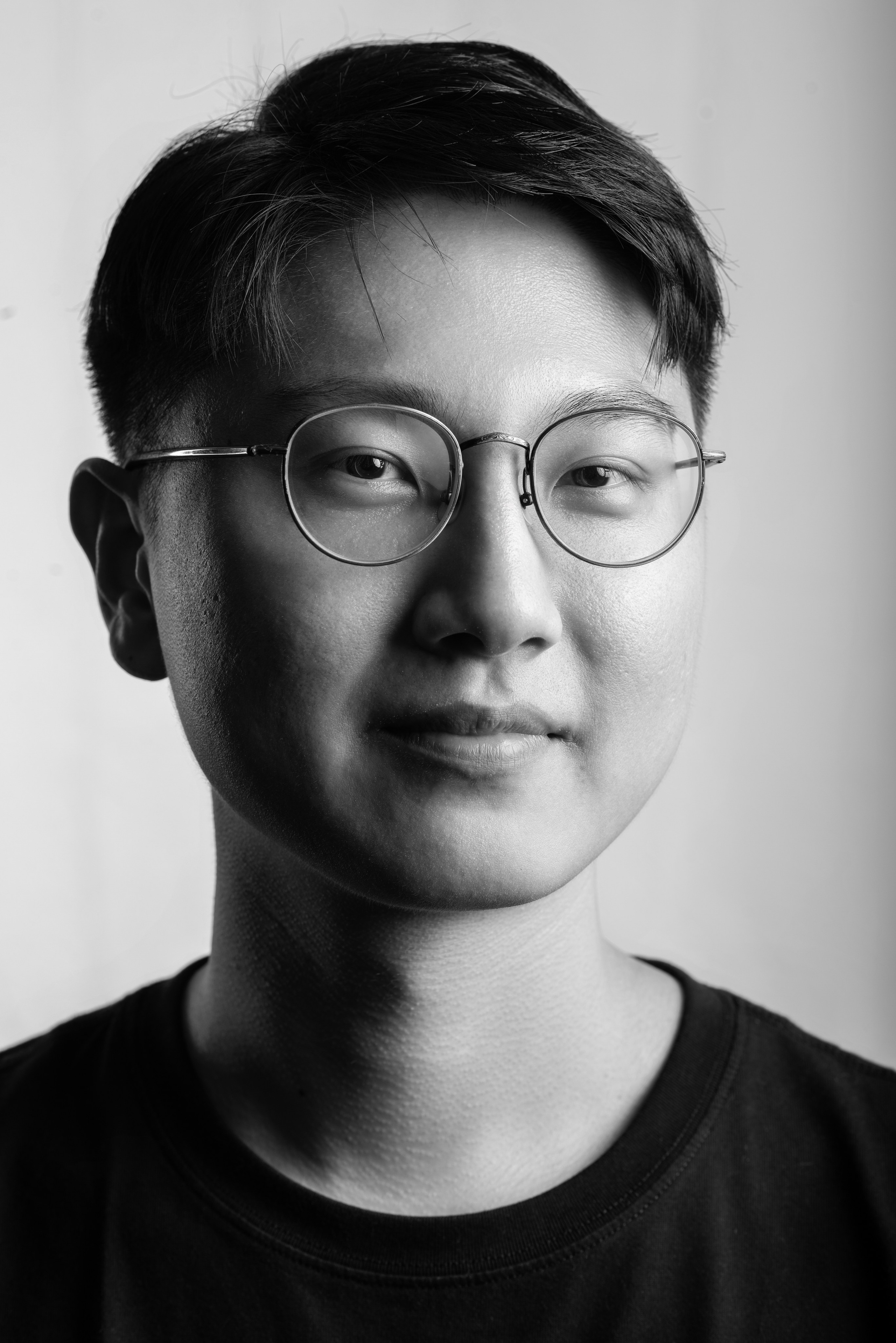 A clean and professional black-and-white corporate portrait of a young man with short, neatly styled hair, round glasses, and a casual black t-shirt. Photographed with soft lighting against a plain white background, the image highlights the subject's approachable and composed demeanor. Ideal for showcasing modern corporate portrait photography for businesses in Singapore prioritizing authenticity and professionalism.