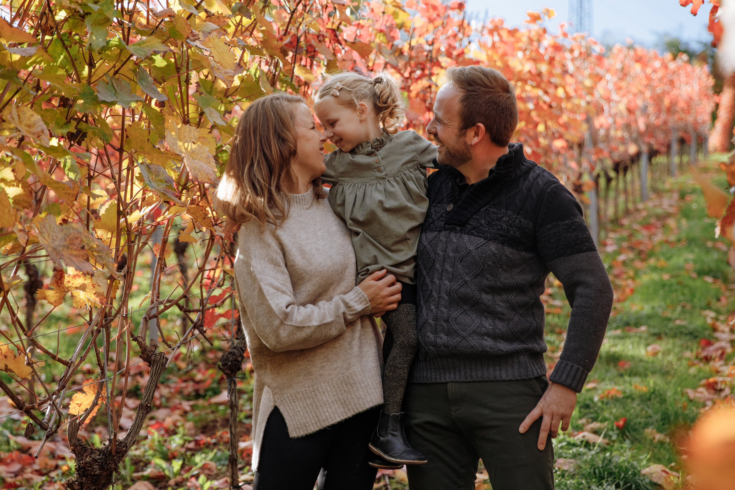 Papa und Mama halten ihre Tochter im Arm in den Weinbergen und lächeln.