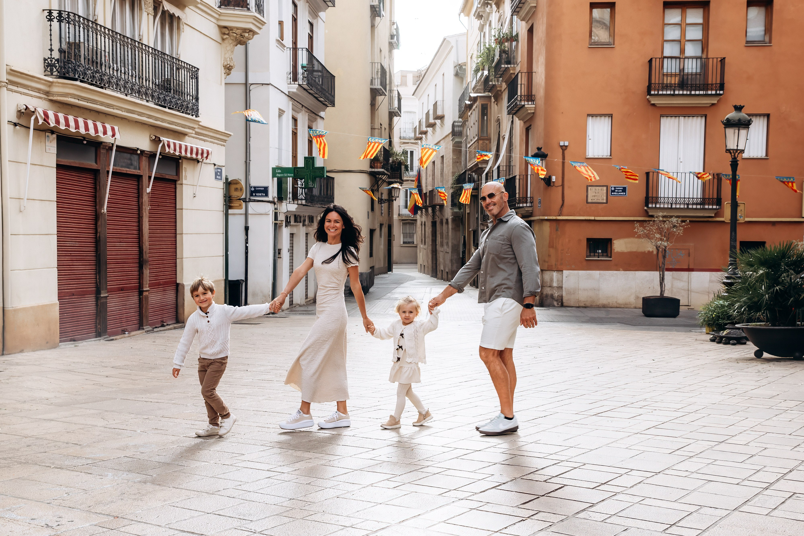 Sesión de fotos familiar en Valencia, España, con una feliz familia de cuatro caminando de la mano por una pintoresca calle del casco histórico de Valencia. Un retrato familiar lifestyle natural que captura sonrisas auténticas, unión y el encanto atemporal del Mediterráneo. Ideal para fotografía familiar, sesiones de fotos familiares, fotografía infantil, retratos familiares de vacaciones y fotografía familiar de destino en Valencia y toda España.