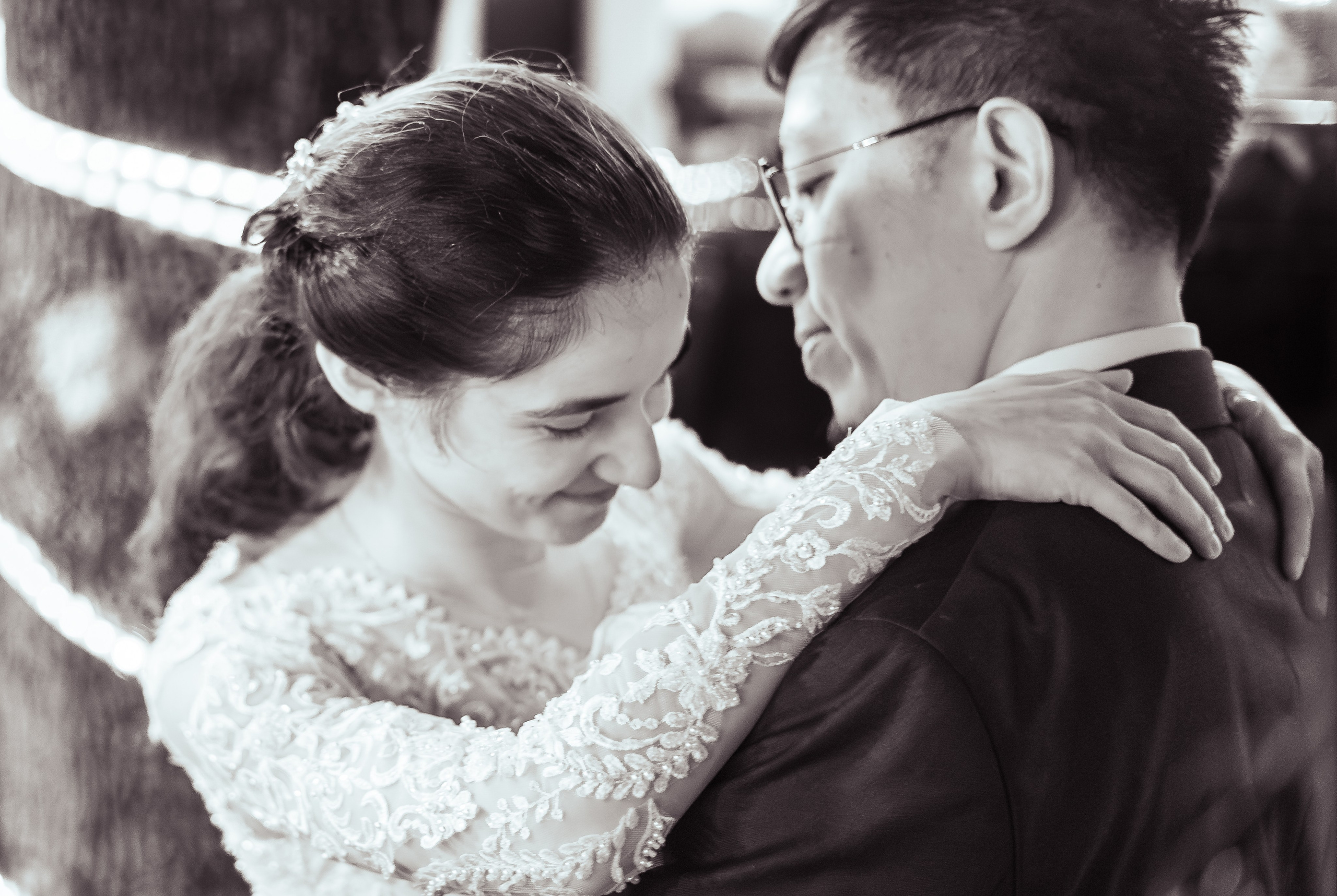 Portret de nuntă alb-negru cu mirii îmbrățișați la primul dans, fundal cu luminițe calde înfășurate pe copac, incadru.ro.