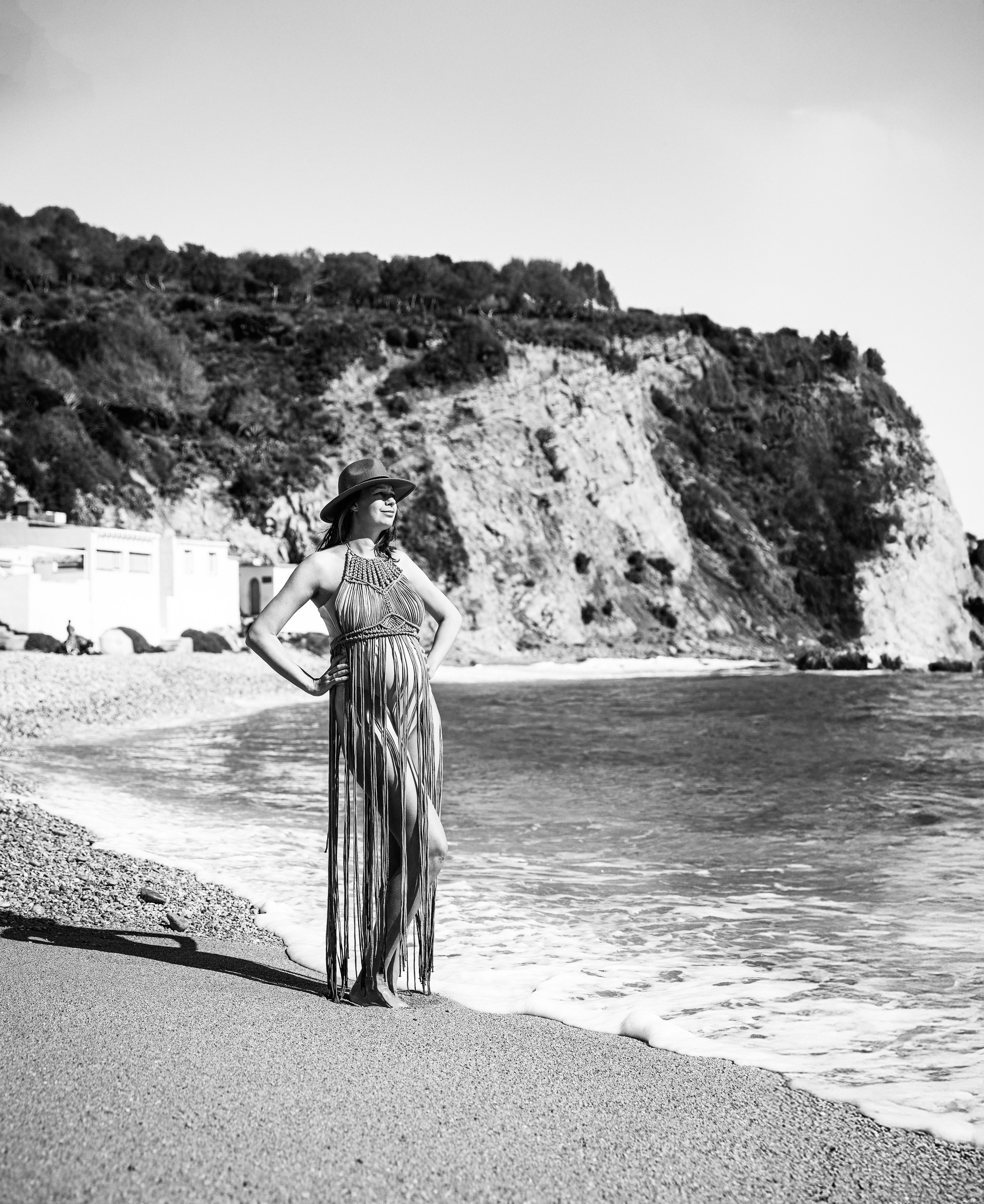 Retrato de maternidad en blanco y negro en Valencia, España — mujer con vestido de flecos y sombrero posando con seguridad al borde del mar con acantilados y olas mediterráneas.