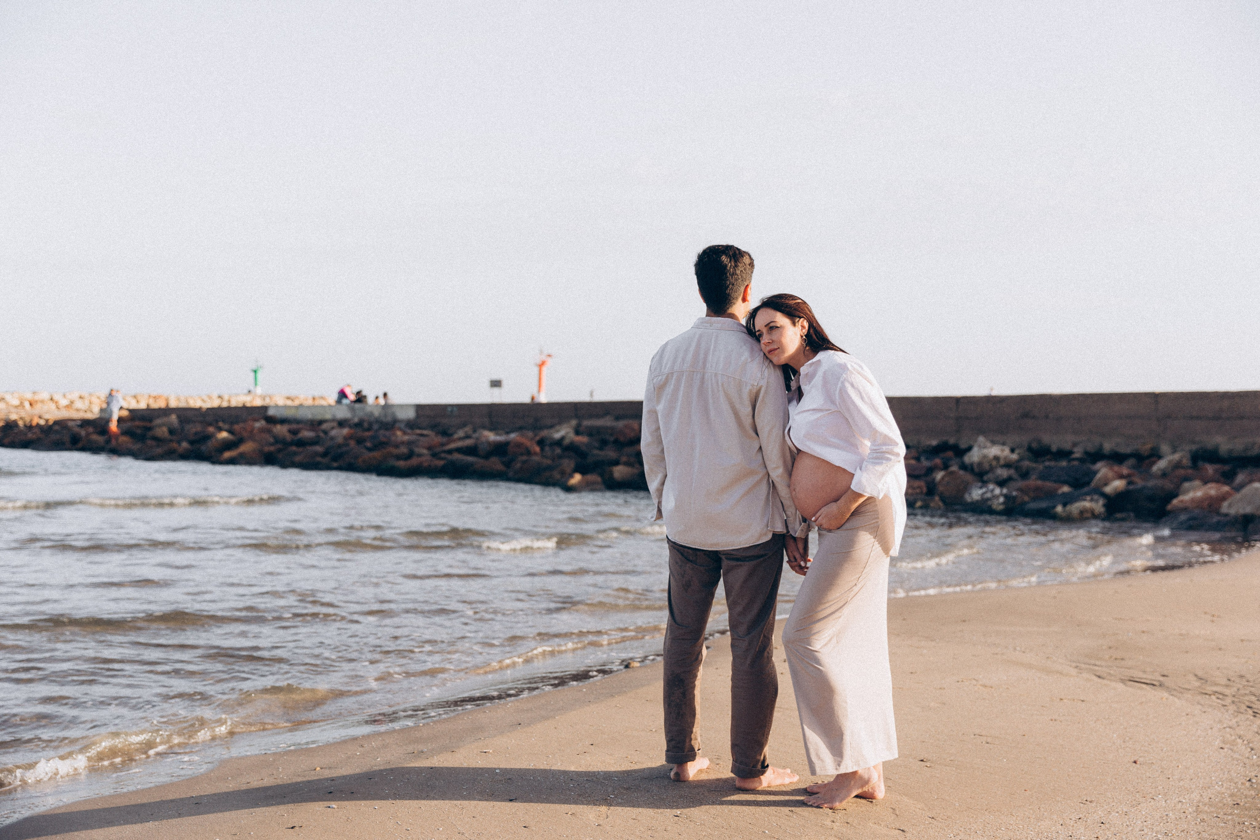 Tierno momento de embarazo capturado en la playa de Valencia, España — mujer embarazada apoyada suavemente sobre su pareja, ambos descalzos cerca de la orilla con las olas suaves y un espigón de fondo.