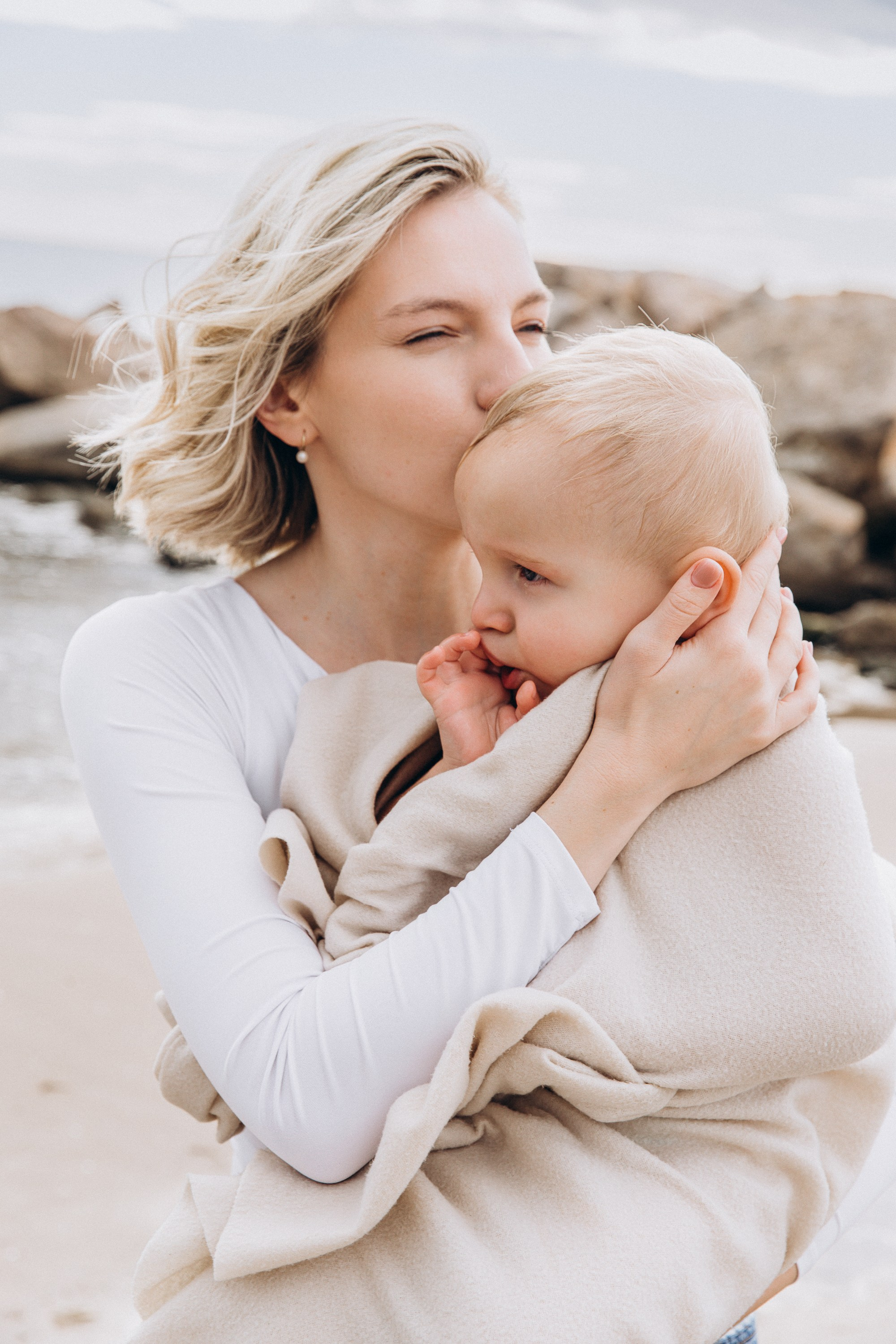 Momento tierno durante una sesión de fotos familiar en Valencia, España — una madre amorosa besa suavemente la cabeza de su bebé mientras lo abraza junto al mar. Un ejemplo perfecto de fotografía familiar natural y emocional en Valencia para quienes buscan sesiones auténticas y conmovedoras en España.