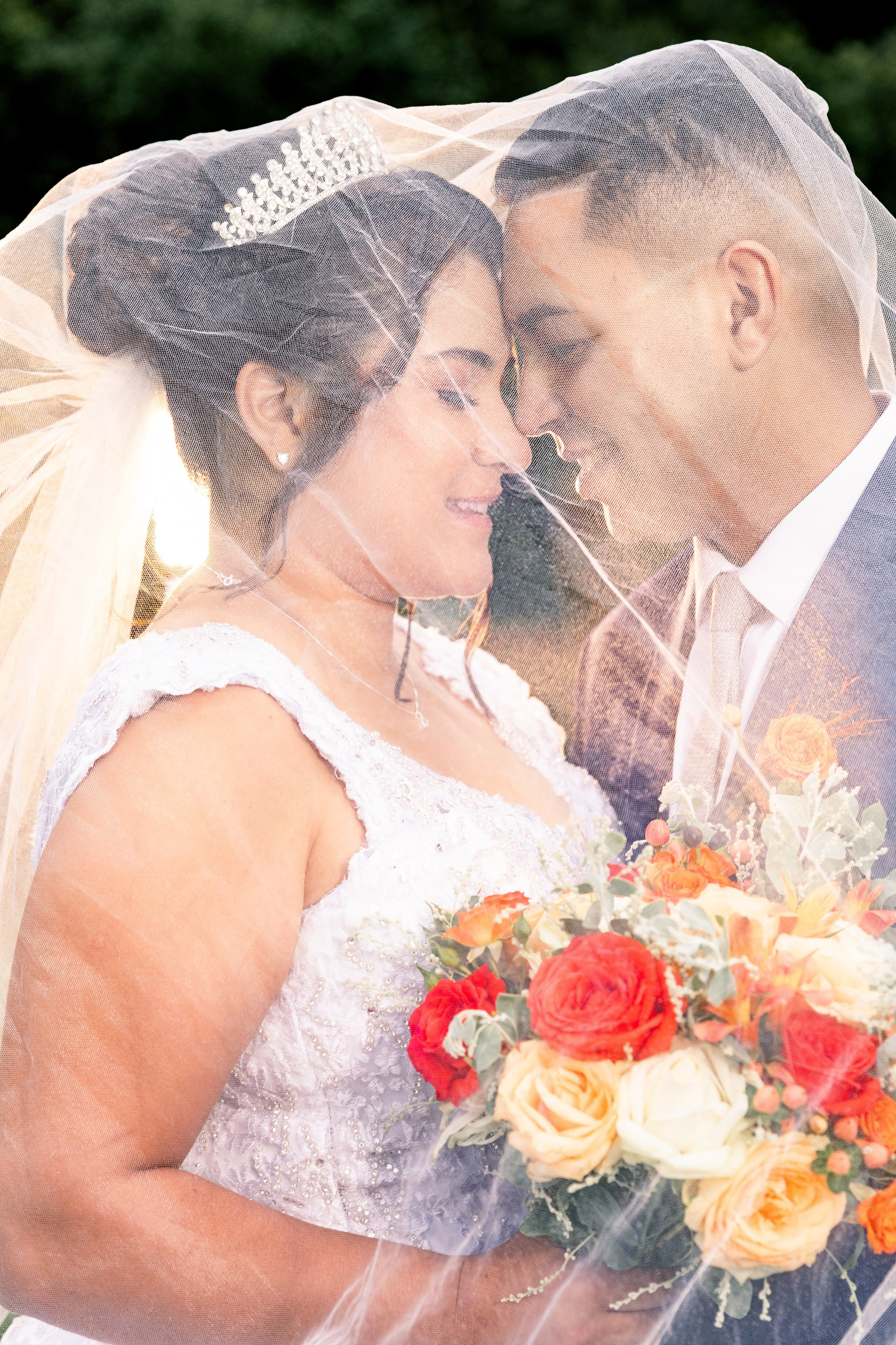 Noivos sob o véu com testas encostadas e olhos fechados, imagem suave e carregada de emoção.