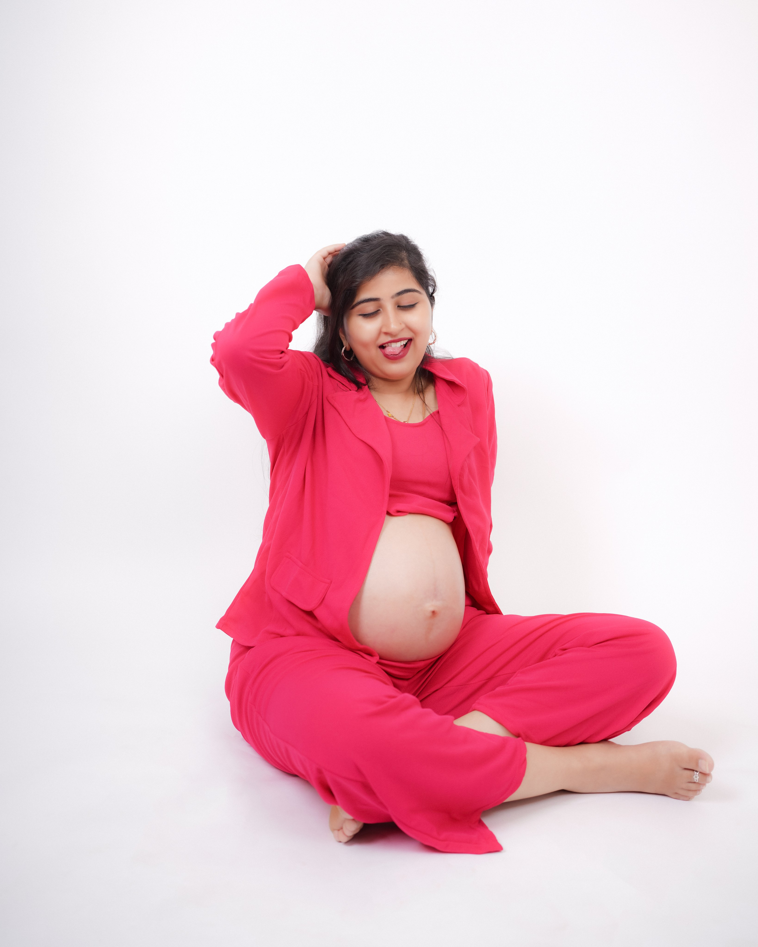 Playful maternity photoshoot in Bengaluru featuring a woman in a pink blazer and pantsuit sitting on a white studio floor, showing her baby bump.