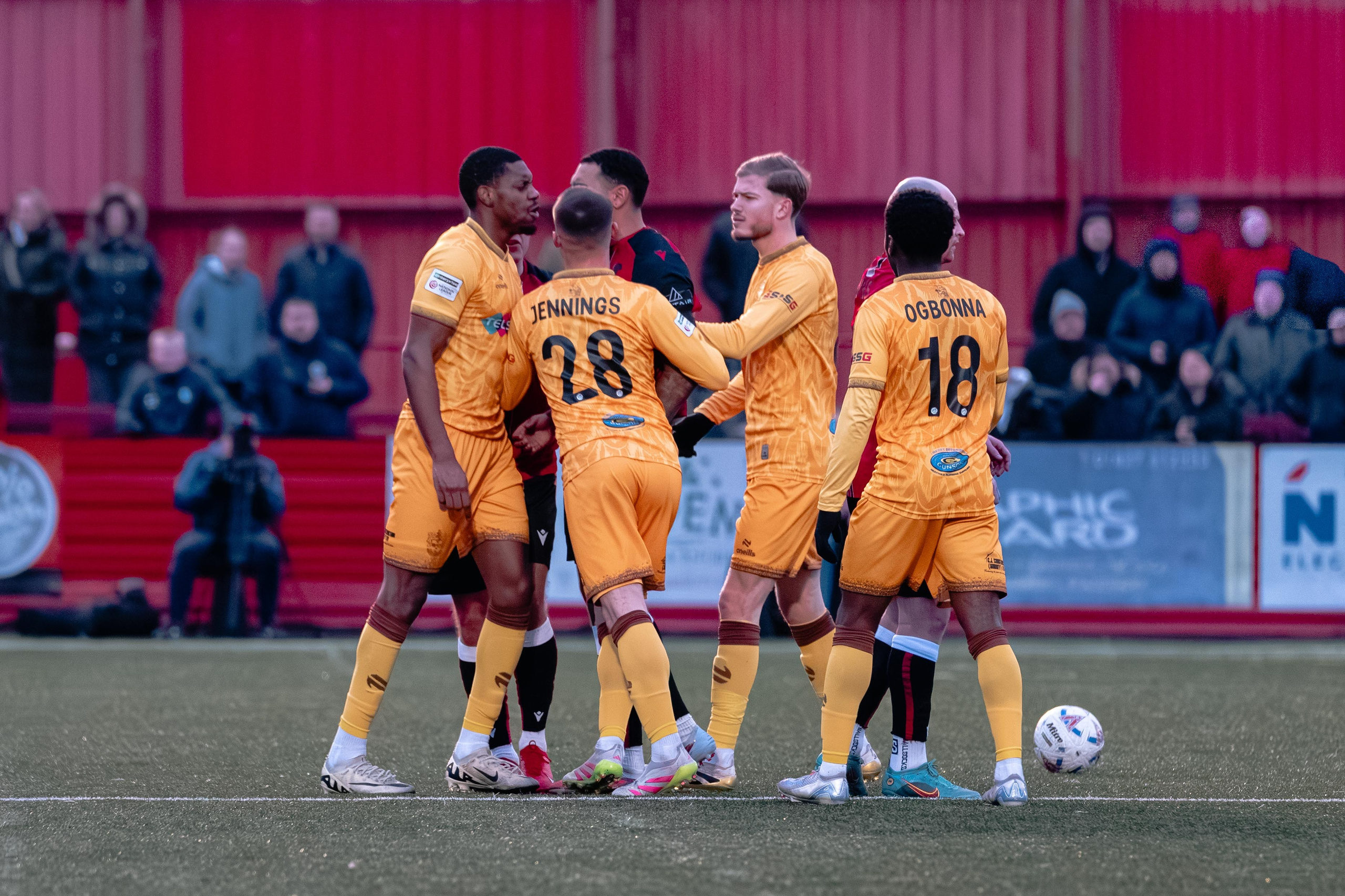Sutton United players in yellow kits gather with Tamworth players during a stoppage, with the ball nearby
