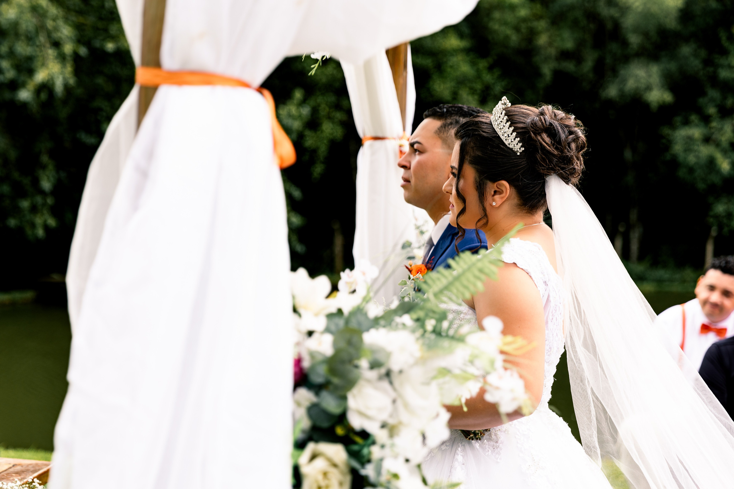 Vista lateral dos noivos no altar com flores em destaque no primeiro plano, momento de cumplicidade.