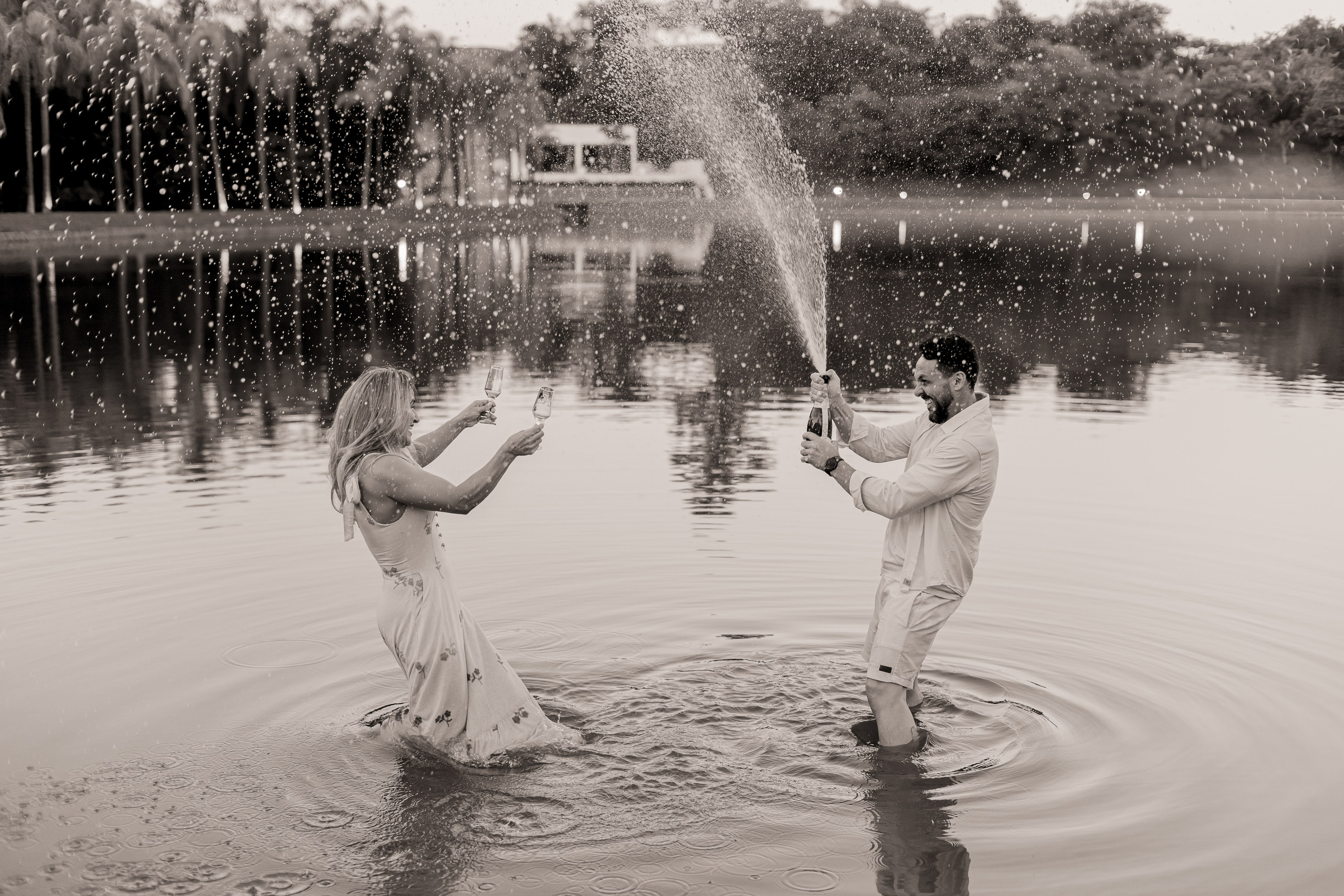 Casal celebrando com champanhe dentro do lago no Resort das Oliveiras ao entardecer, com espirros d'água e sorrisos
