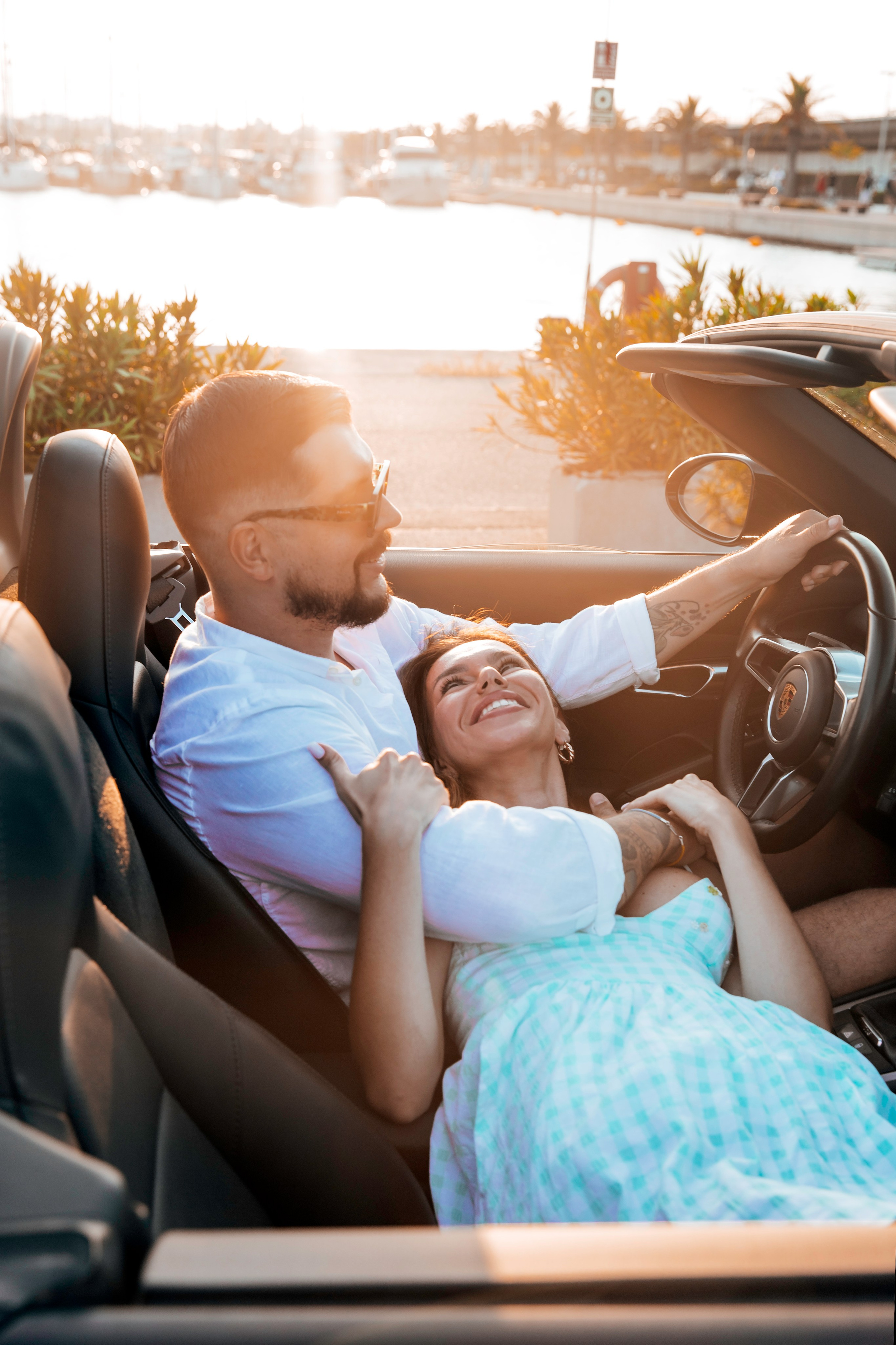 A couple shares a joyful and intimate moment in a convertible at sunset, parked along a marina in Valencia, Spain, with golden light illuminating their smiles and the calm water in the background.