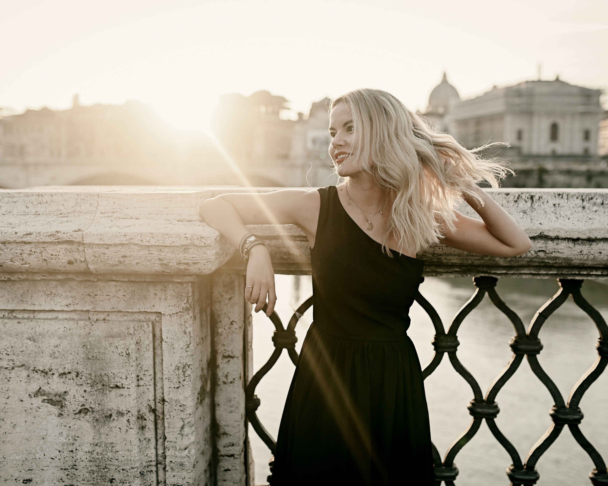 Woman in a black dress smiles, leaning on a marble railing above the Tiber River at sunset