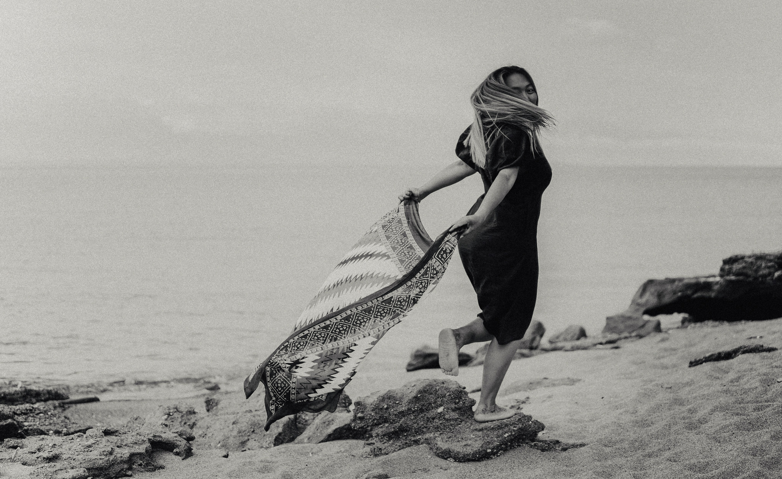 На черно-белой фотографии изображена девушка, бежащая вперёд по побережью моря. Ее яркий шёлковый платок развевается за ней, создавая атмосферу движения и свободы.