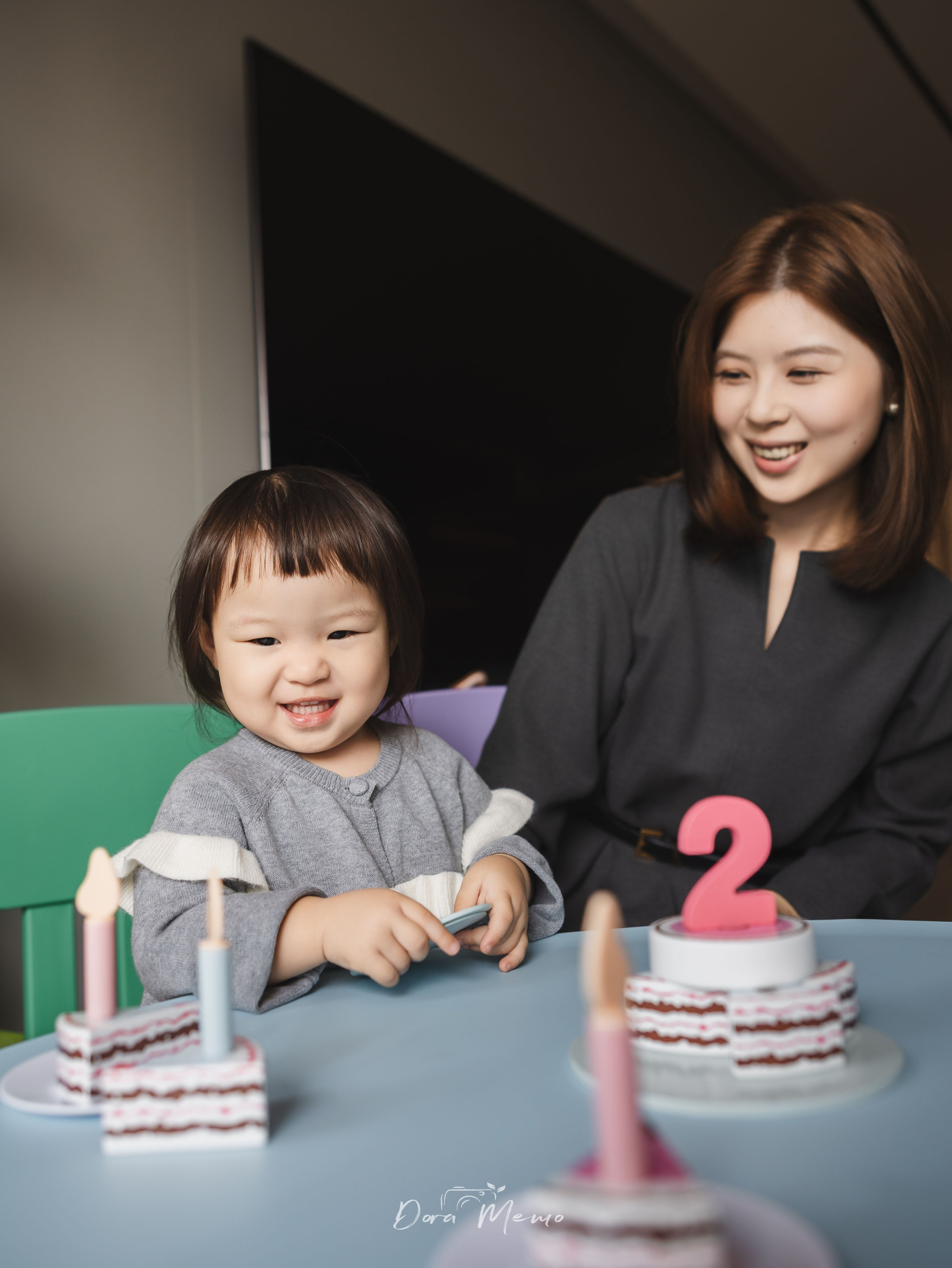 Two-year-old birthday girl sitting at a table with a small cake and number 2 candle, milestone family photography at home.