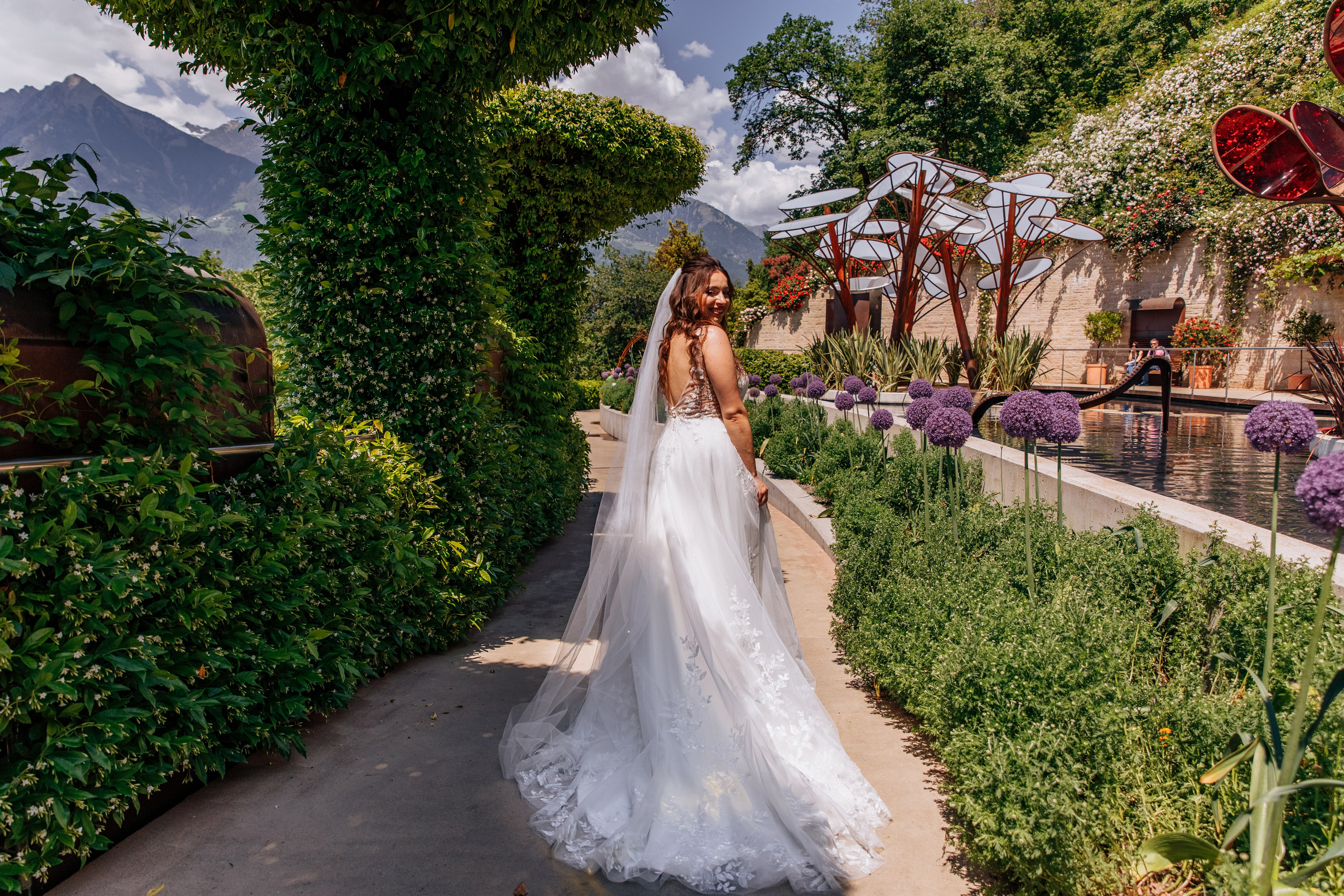 Die Braut geht mit ihrem wunderschönen Hochzeitskleid und einem eleganten Schlepptail durch einen blühenden Garten, den Rücken zu uns gewandt