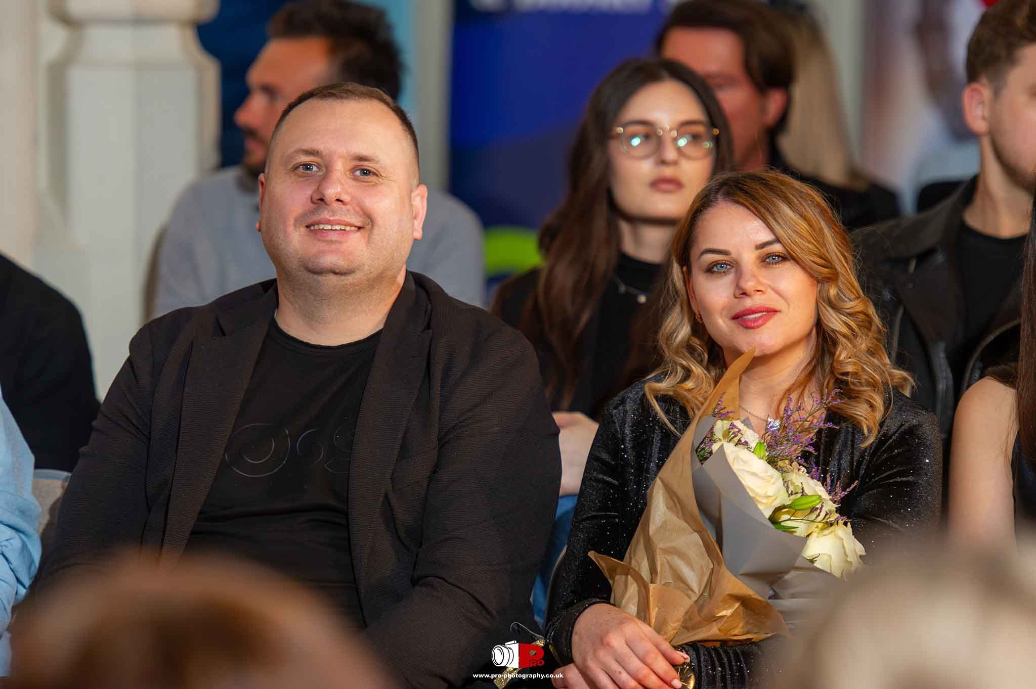 A smiling man and woman are attending an award ceremony, with the woman holding a bouquet of white roses.