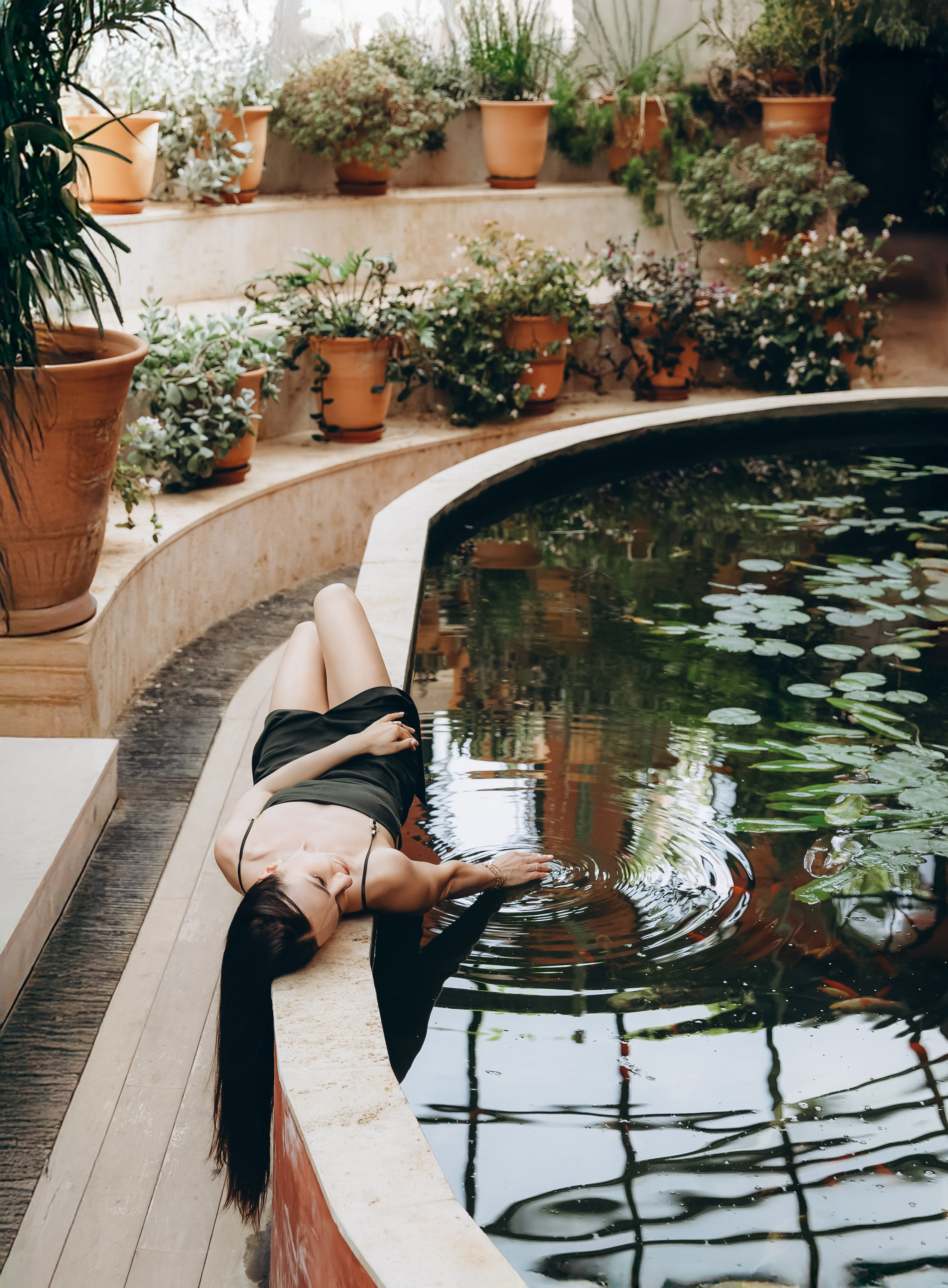 Женщина в черном платье лежит у пруда с лилиями и касается воды, окружённая терракотовыми горшками с растениями в залитом солнцем зимнем саду в Барселоне, Испания — спокойная и креативная портретная фотография, сочетающая элегантность, природу и средиземноморское очарование, идеально подходит для портретных фотосессий в Барселоне и Испании.