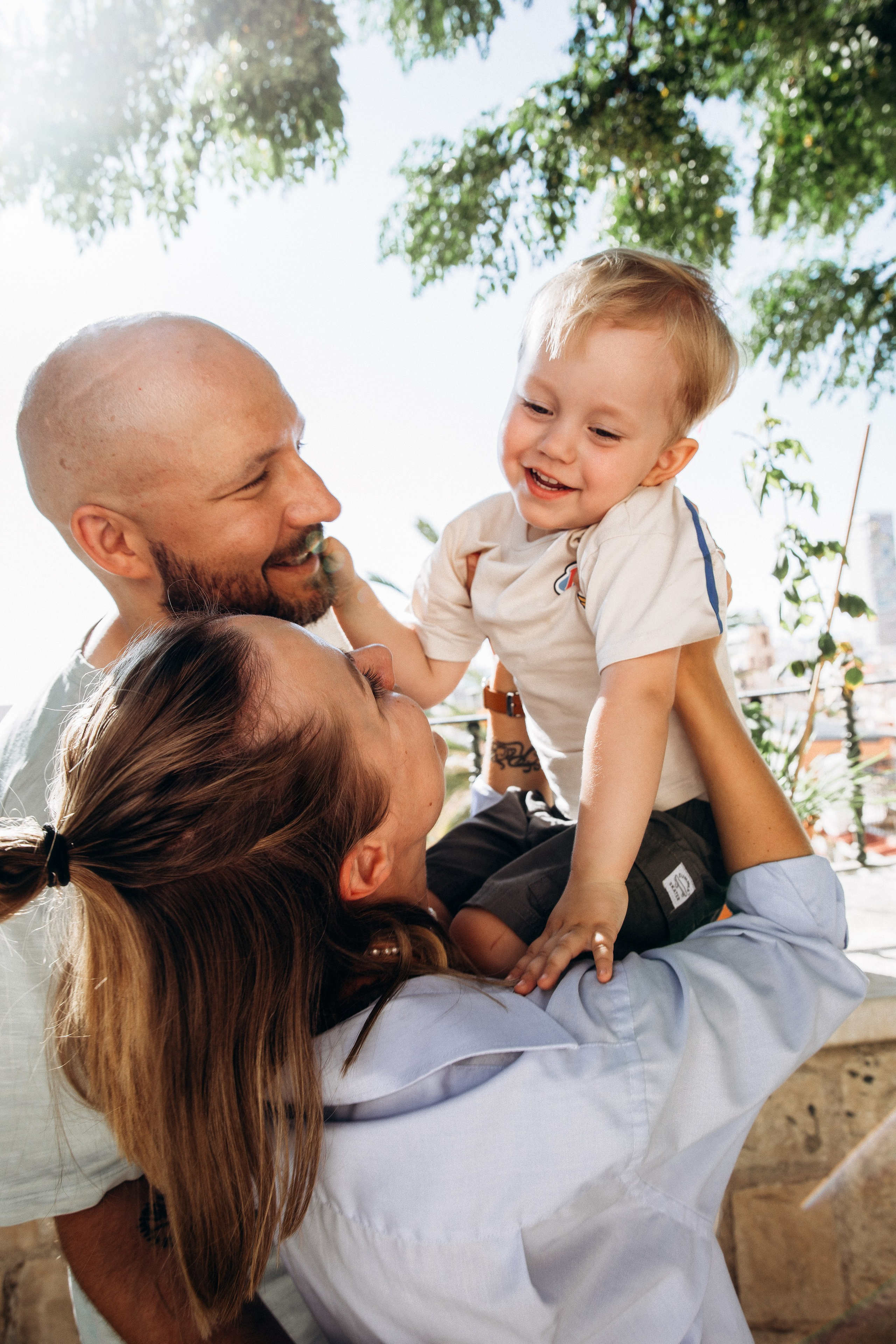 Momento familiar alegre capturado en Valencia, España, mostrando a una pareja sonriente levantando a su hijo feliz bajo árboles frondosos — ejemplo perfecto de sesiones familiares naturales y espontáneas en Valencia y en toda España.