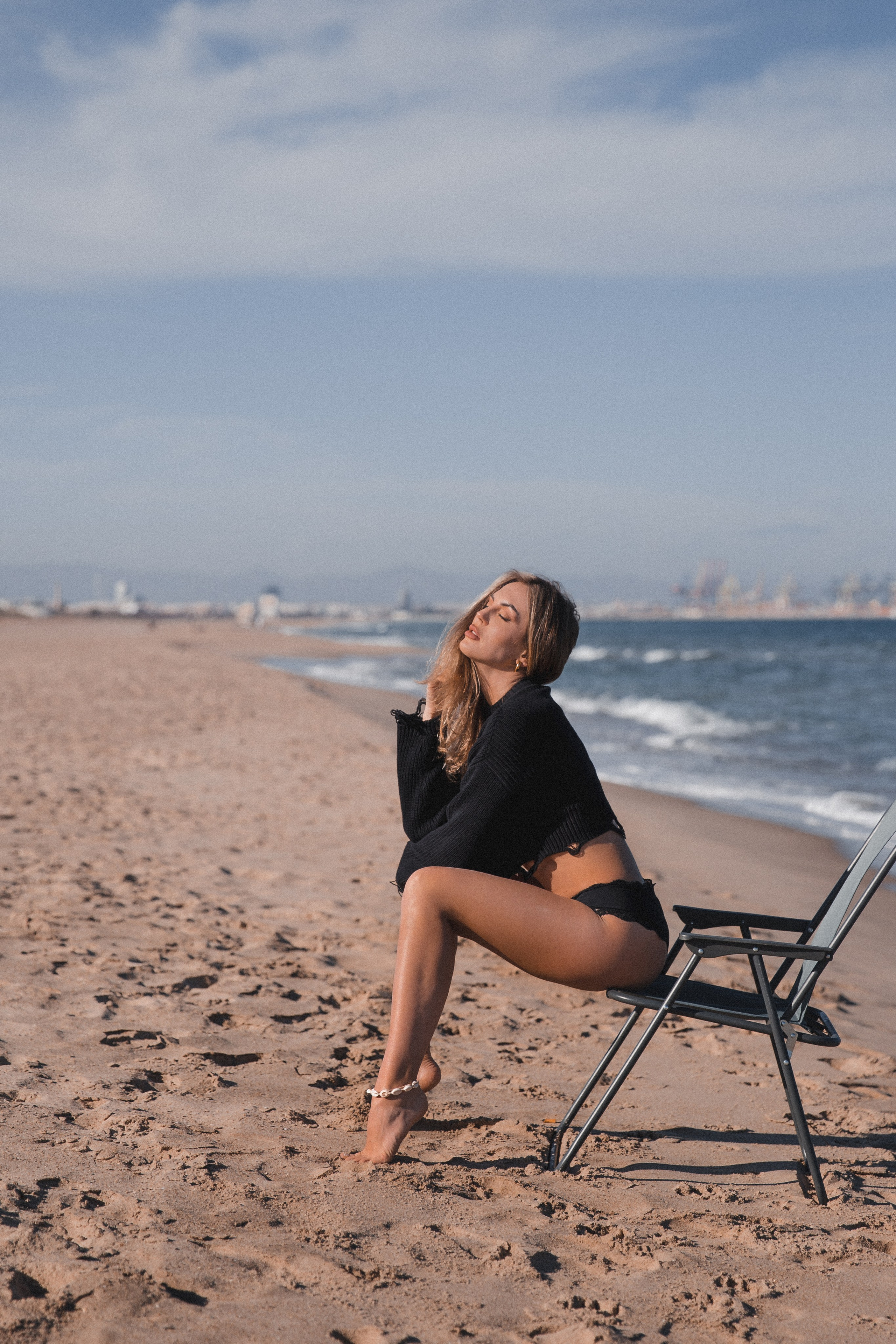 Модный портрет женщины в черном свитере и нижней части бикини, сидящей на стуле, уверенно позирующей на песчаном пляже с морем и городским горизонтом на заднем плане, в Валенсии, Испания