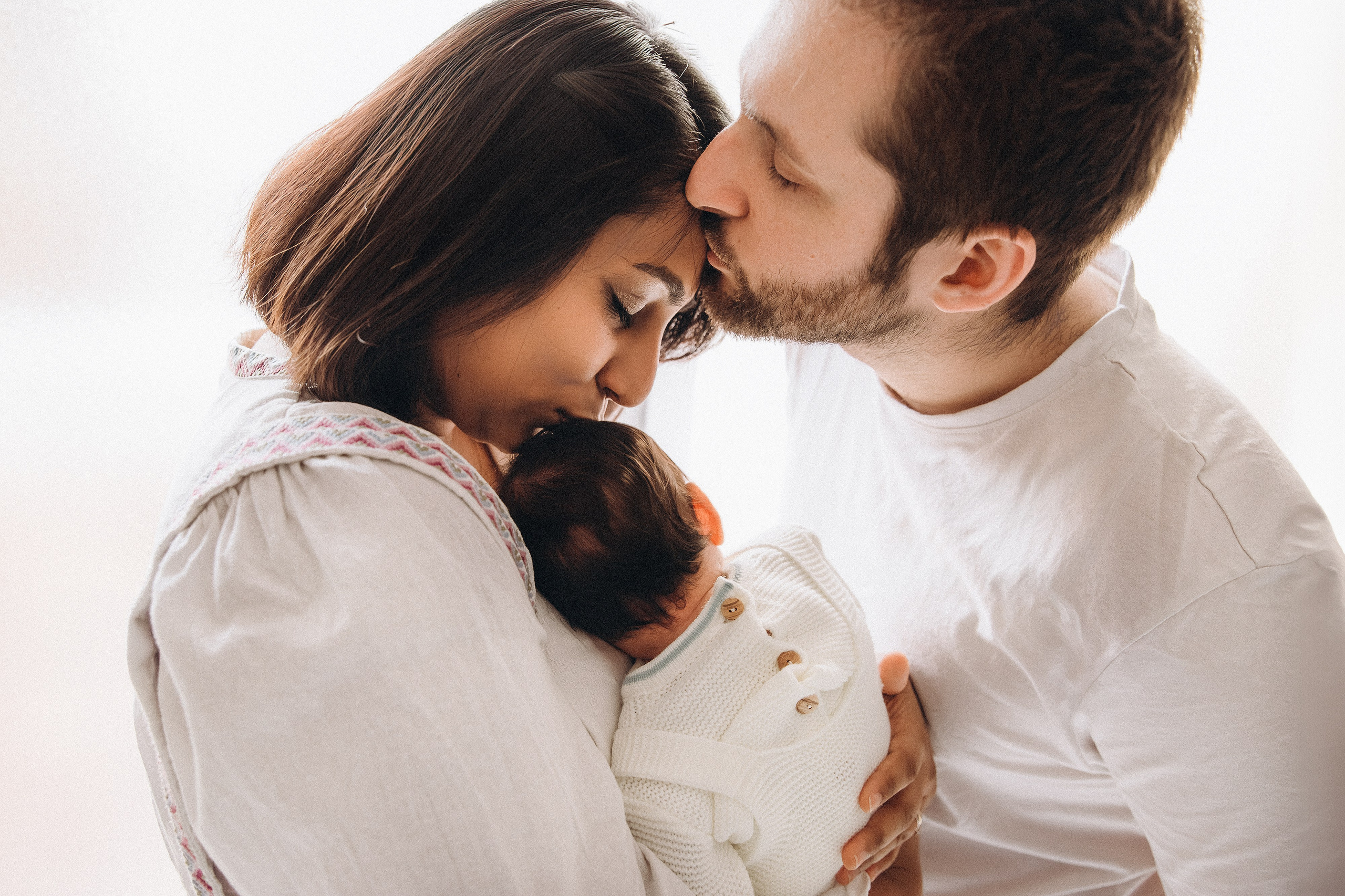 Семейная фотосессия в Валенсии, Испания, где родители держат своего новорождённого малыша и делятся нежным поцелуем, снято в мягком естественном свете для тёплого, эмоционального и интимного семейного портрета — идеально для newborn и семейной фотографии в Валенсии и по всей Испании.