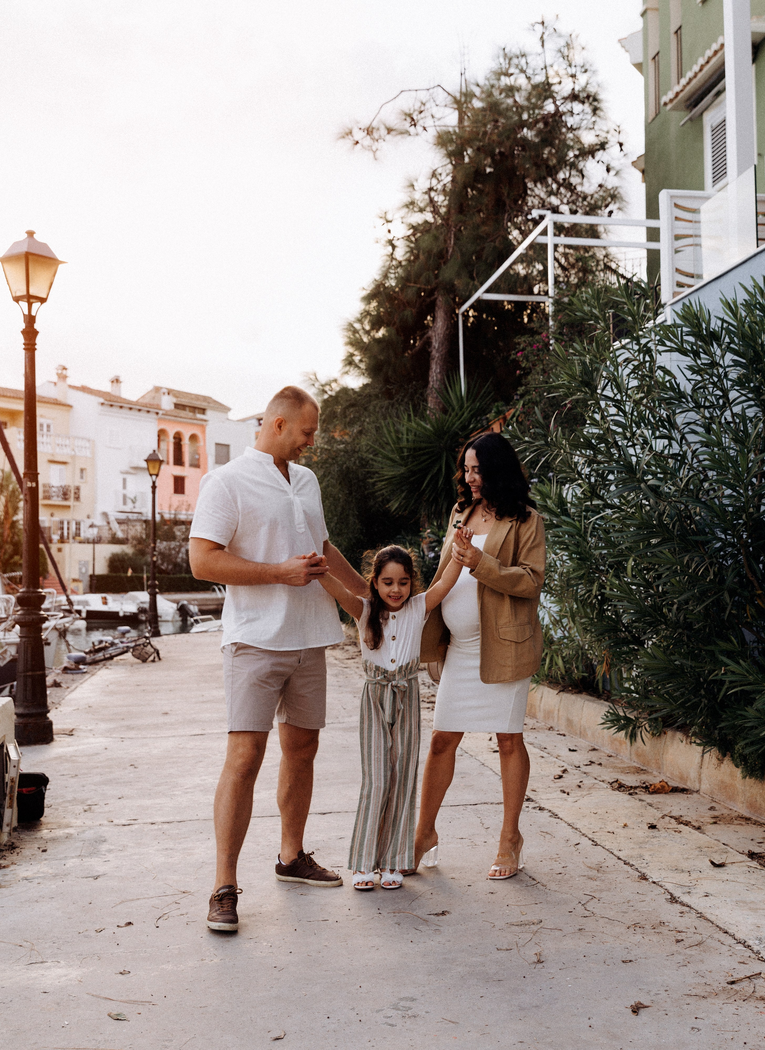 Paseo familiar alegre por las encantadoras calles de Port Saplaya, Valencia, España — madre embarazada, padre e hija compartiendo sonrisas durante la hora dorada.