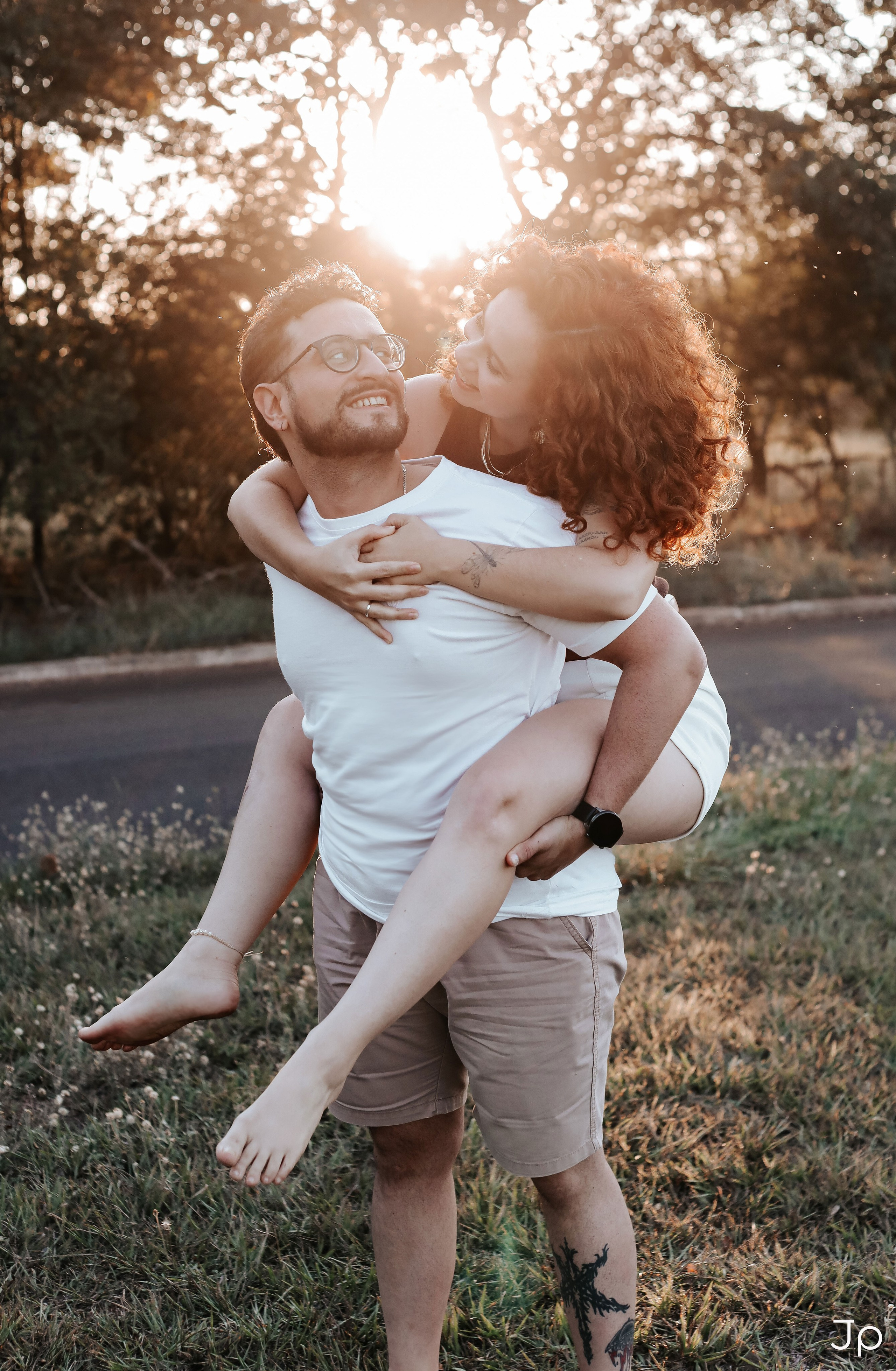 Casal sorrindo, com ela nas costas dele, ao pôr do sol em um ensaio fotográfico.