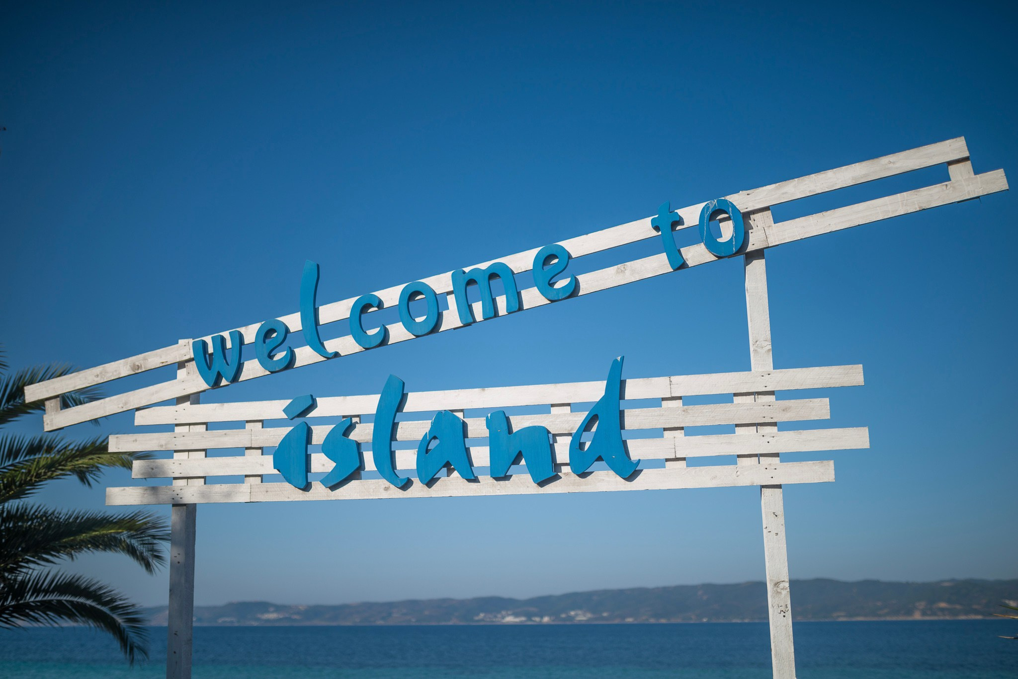 Beli drveni znak „Welcome to Island“ sa plavim slovima, postavljen naspram svetloplavog neba. Ispod se nalaze miran okean i daleka obala.