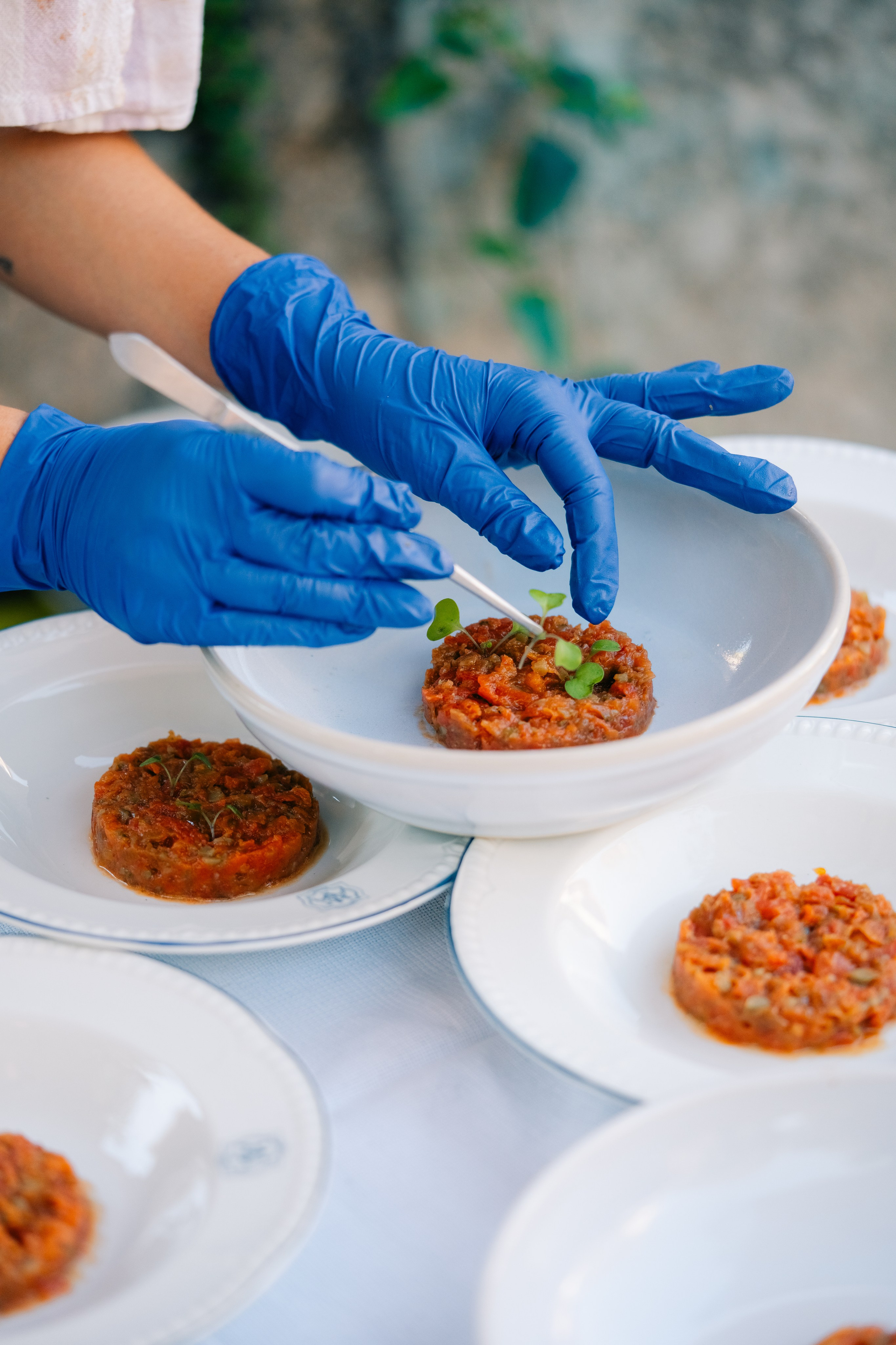Шеф-повар в синих перчатках раскладывает овощной тартар. Шеф-повар в синих перчатках аккуратно раскладывает овощной тартар на белых блюдах, украшая его микрозеленью для изысканной презентации на открытом воздухе.