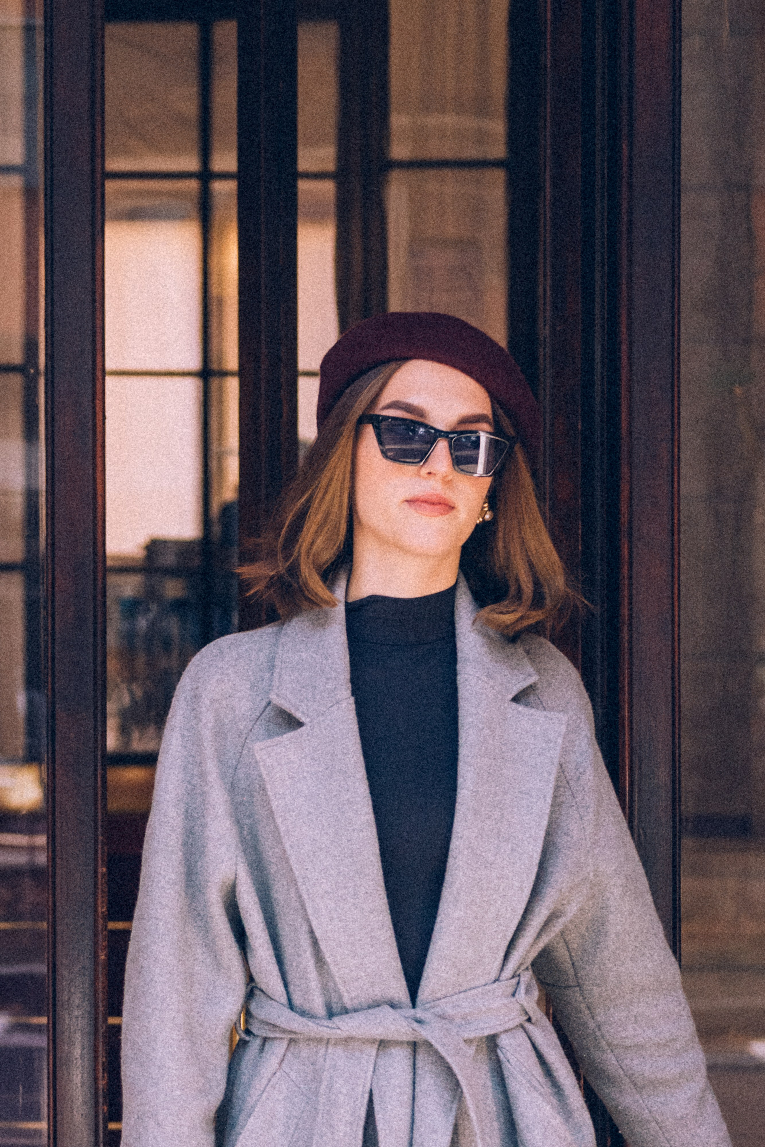 Elegant woman in a gray belted coat and burgundy beret posing in front of a polished glass doorway, showcasing classic Milanese style. Milan photographer for tourists