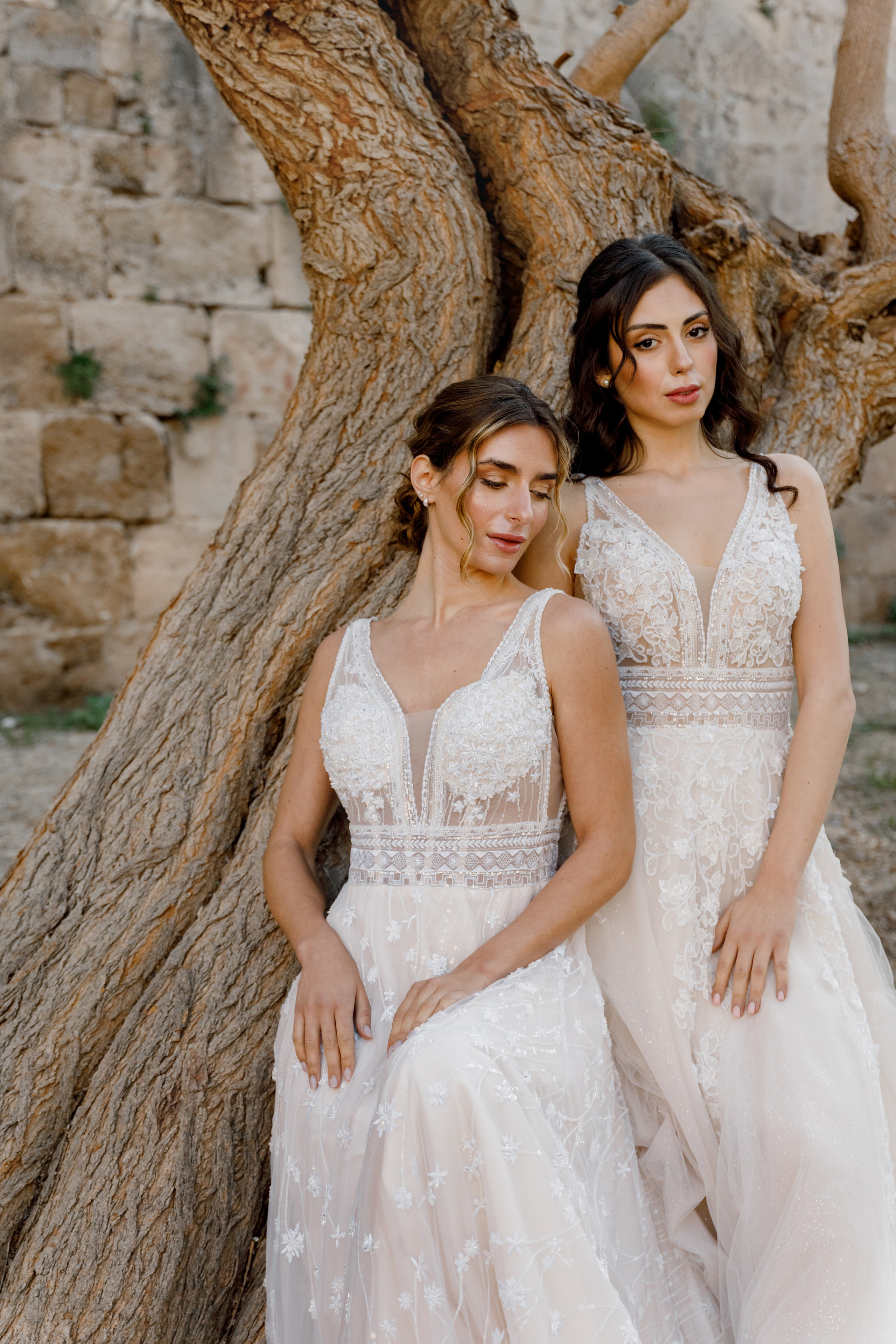 Two brides in elegant wedding dresses pose in the narrow, cobblestone streets of Rhodes' Old Town, surrounded by historic stone buildings and vibrant bougainvillea. The editorial-style photo captures their intimate connection and the timeless charm of the medieval setting, with soft natural light enhancing the romantic atmosphere.