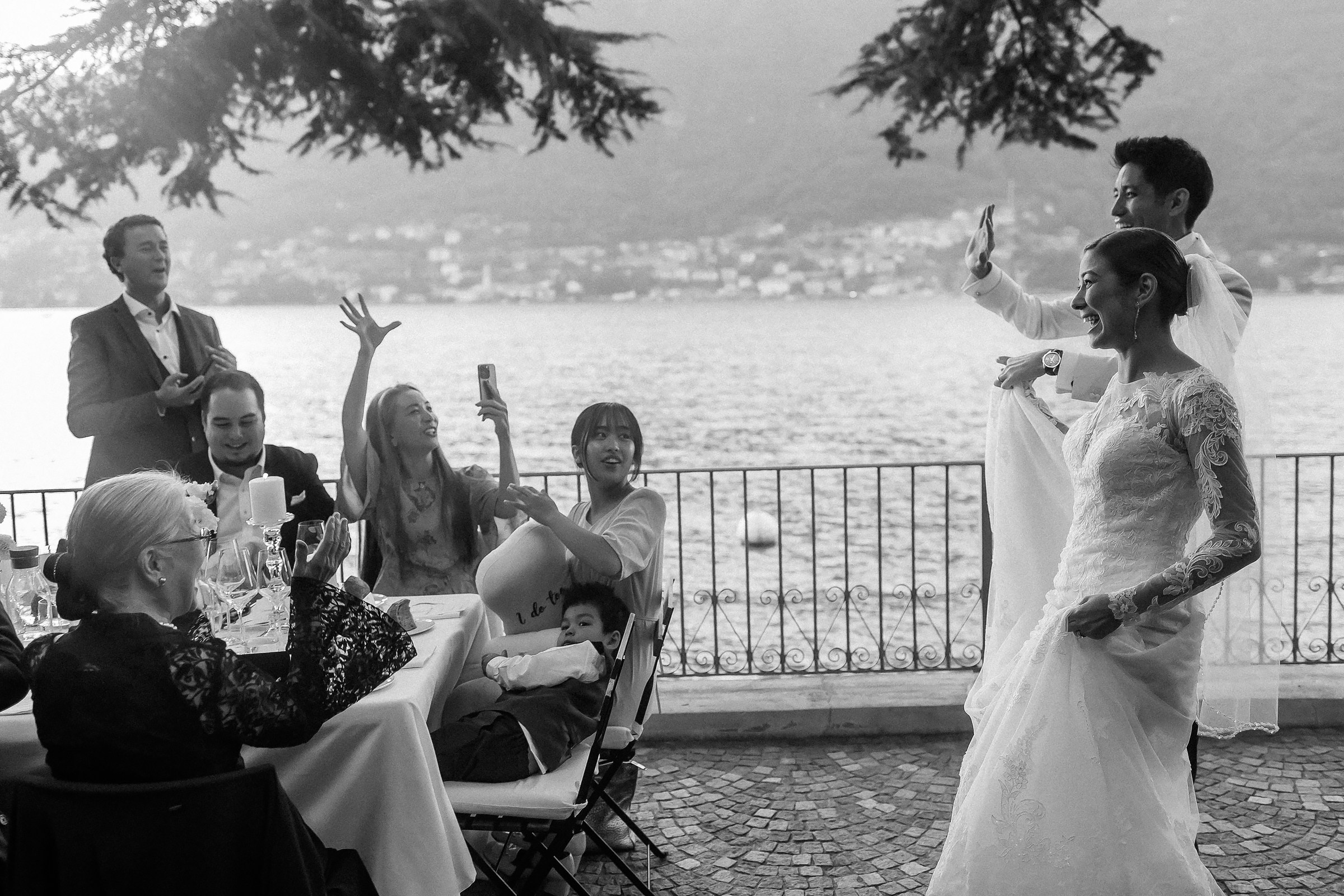 Bride and groom dance joyfully, surrounded by cheering guests at a lakeside table, capturing a festive moment.