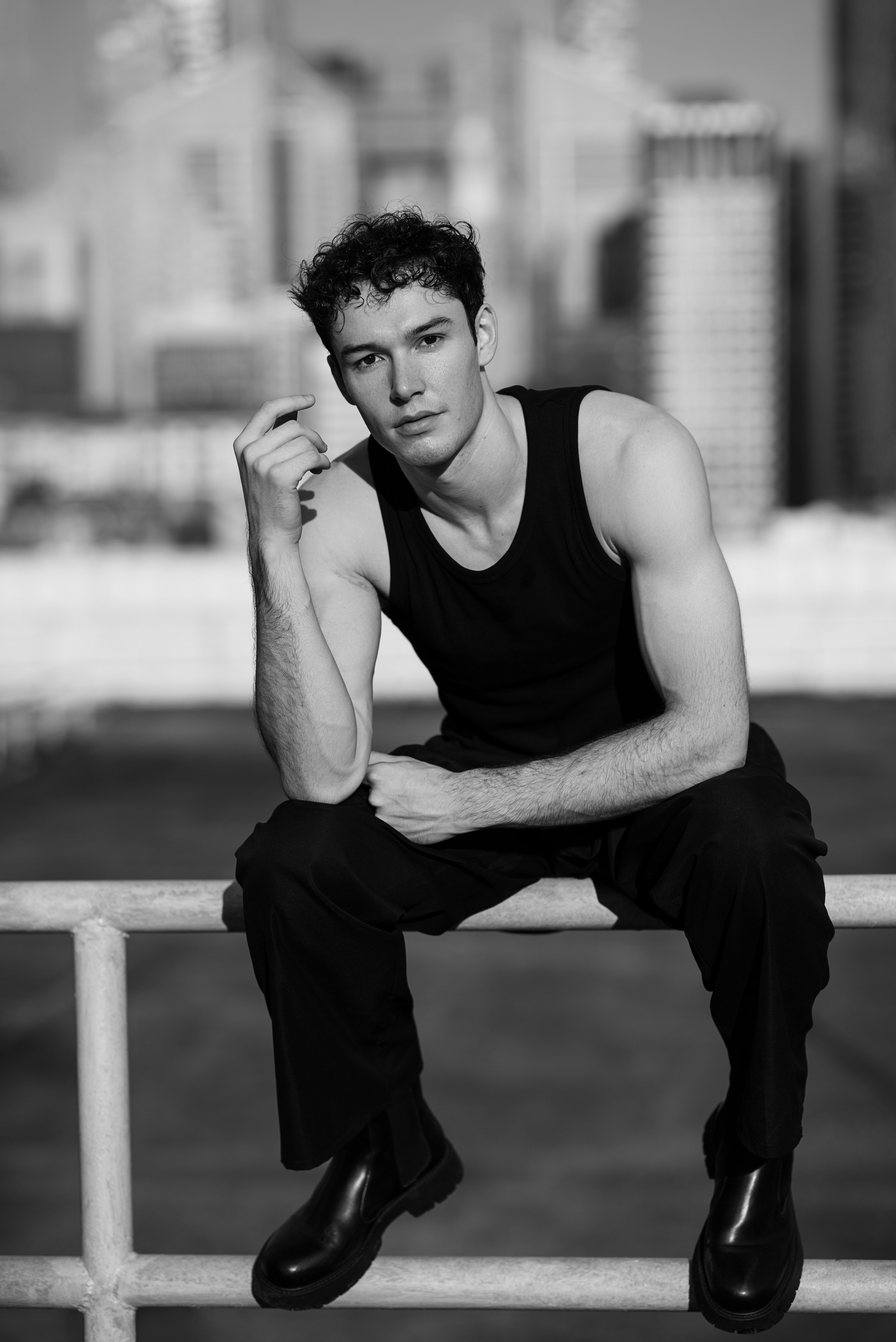 Seated rooftop portrait of a male model in black tank top, city towers of Singapore blurred in the background.