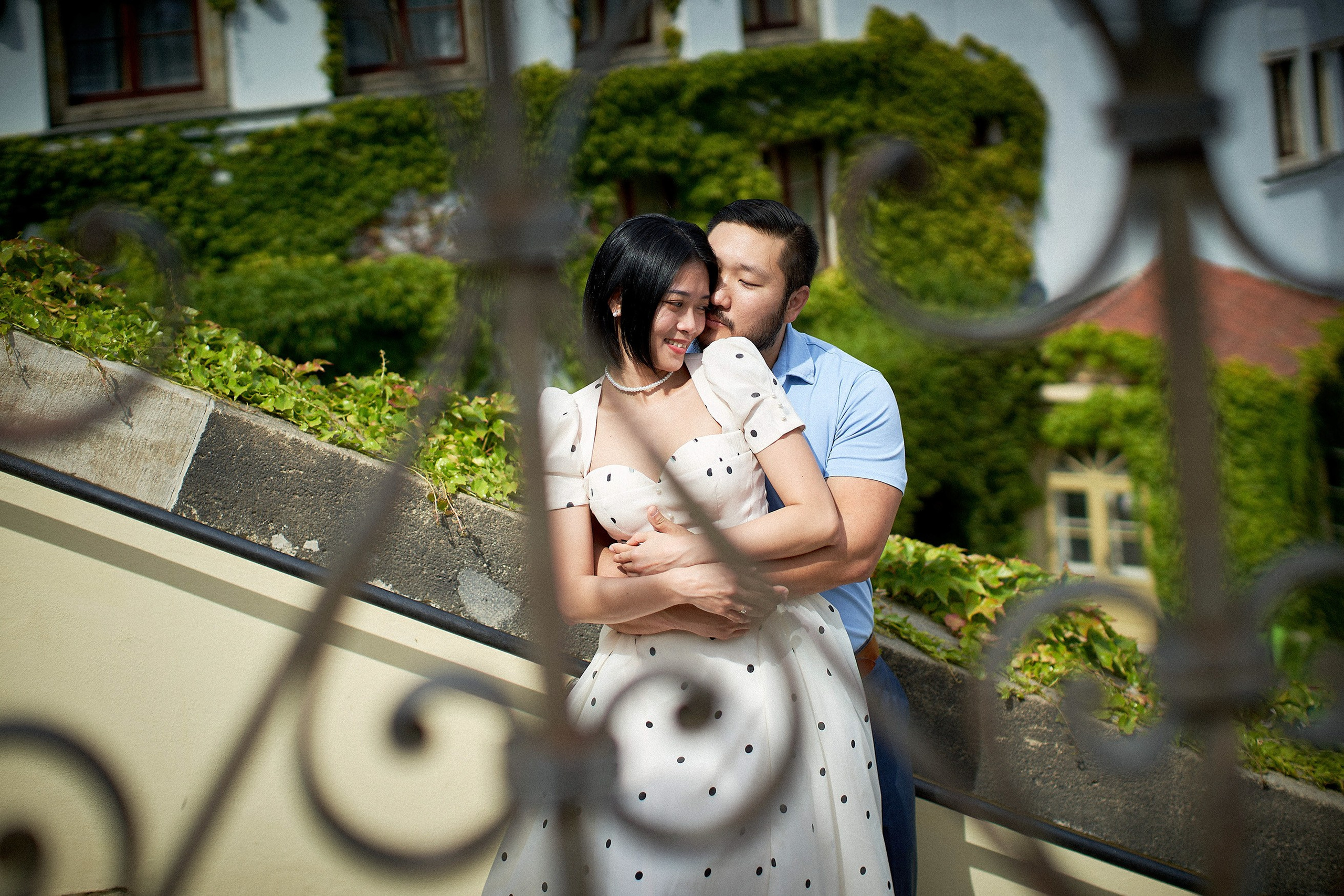 Engaged couple embracing as seen through a decorative wrought-iron railing in Vrtba Garden.