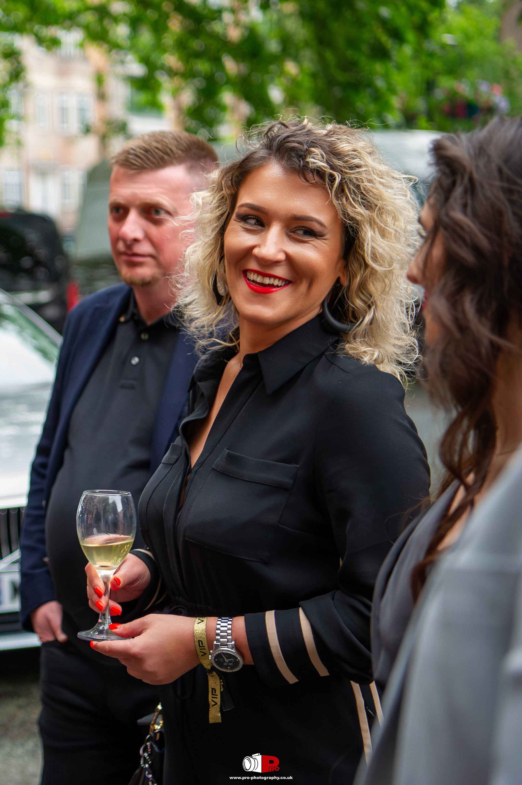 Cheerful woman in black attire holding a wine glass while engaging in a lively outdoor conversation at a social event.