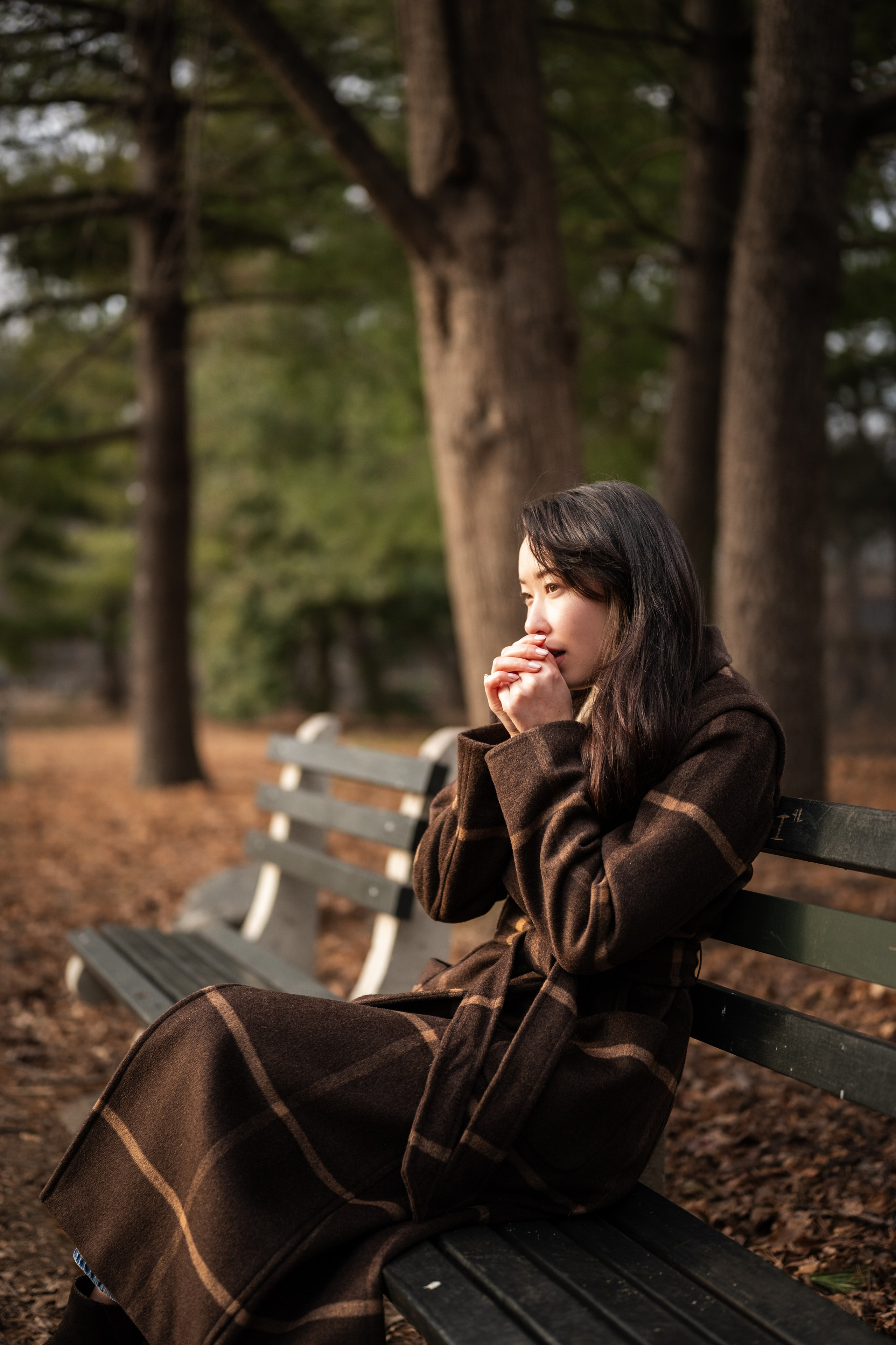 Woman sitting on a bench wrapped in a coat in Van Cortlandt Park, Bronx, New York, cinematic outdoor portrait.