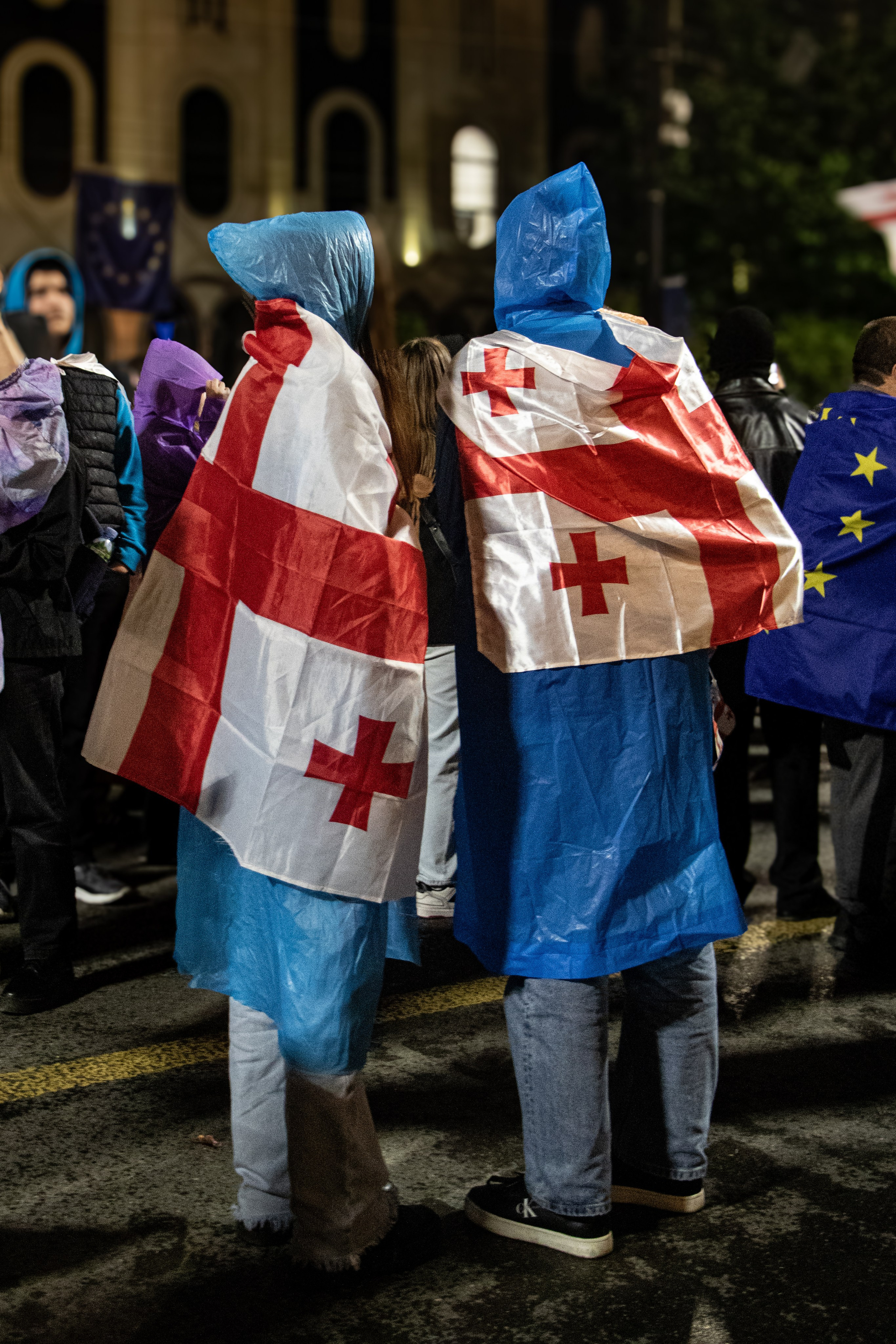 Deux jeunes Géorgiens, drapeaux noués aux épaules, en kway, manifestent devant le parlement, exprimant leur soutien à l’opposition et leur engagement politique dans un contexte tendu à l’approche des élections législatives.
